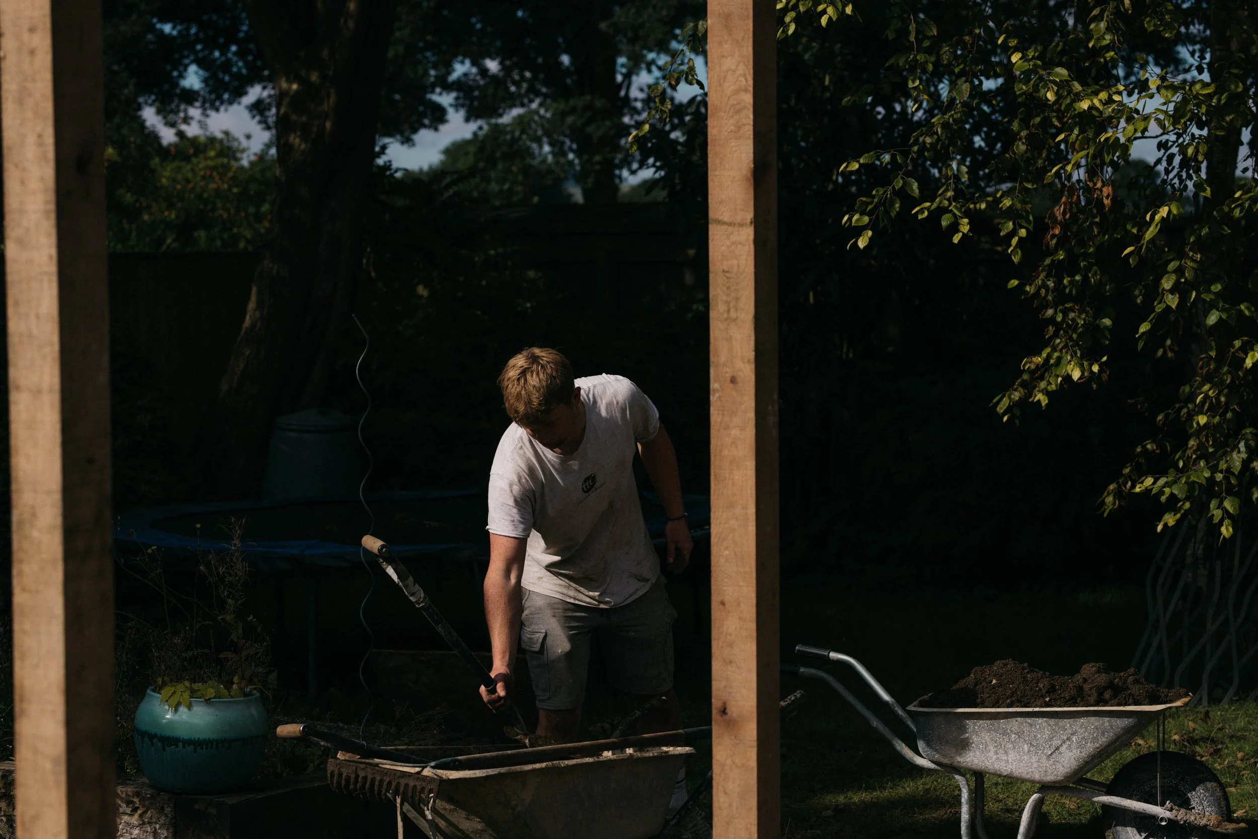 A person working on gardening or construction in a backyard during the evening or early night, with trees and wooden posts visible.