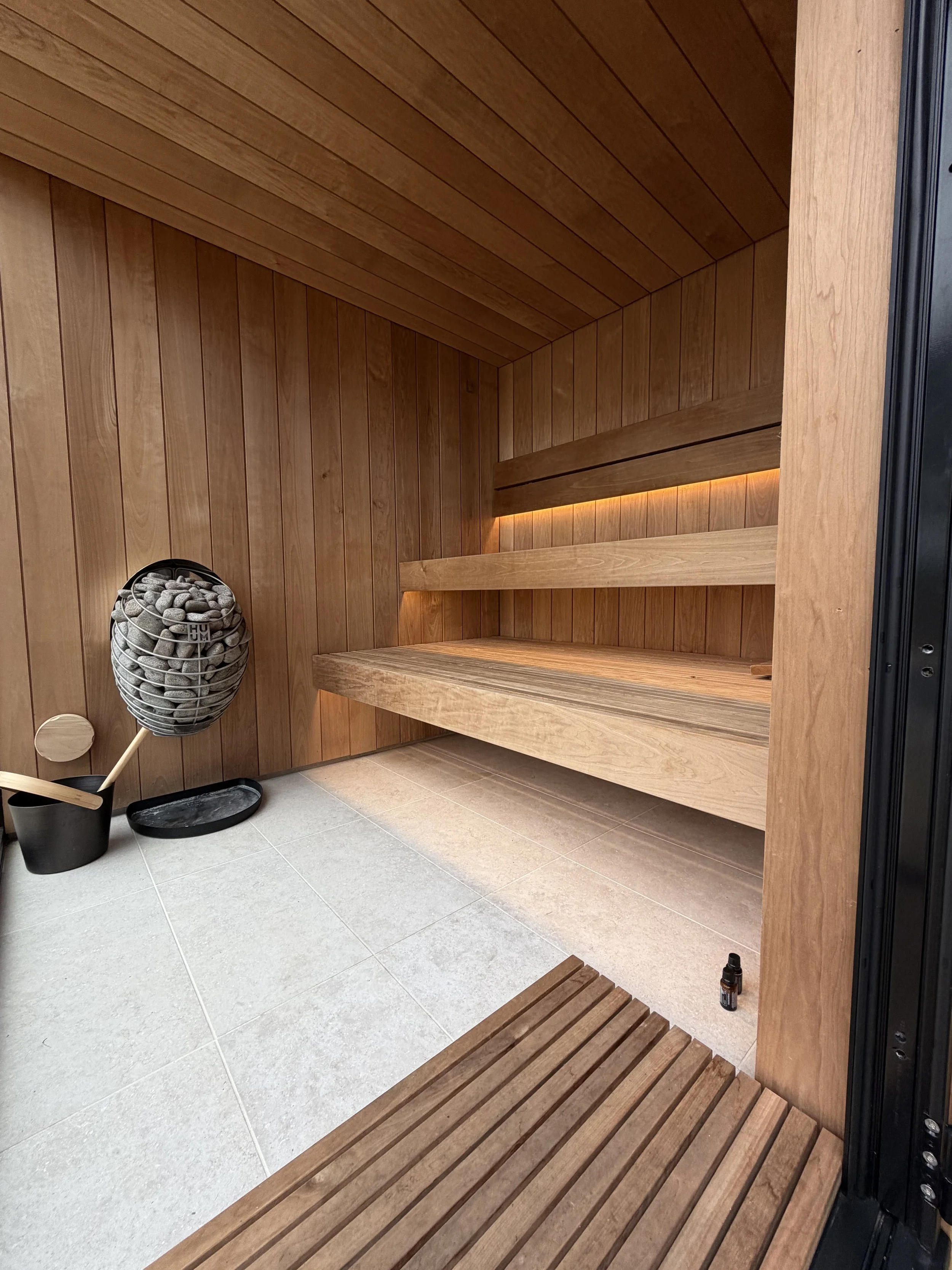 Interior of a wooden sauna with benches, a small black bucket, a wooden ladle, a container of sauna stones, and a small bottle on the floor.