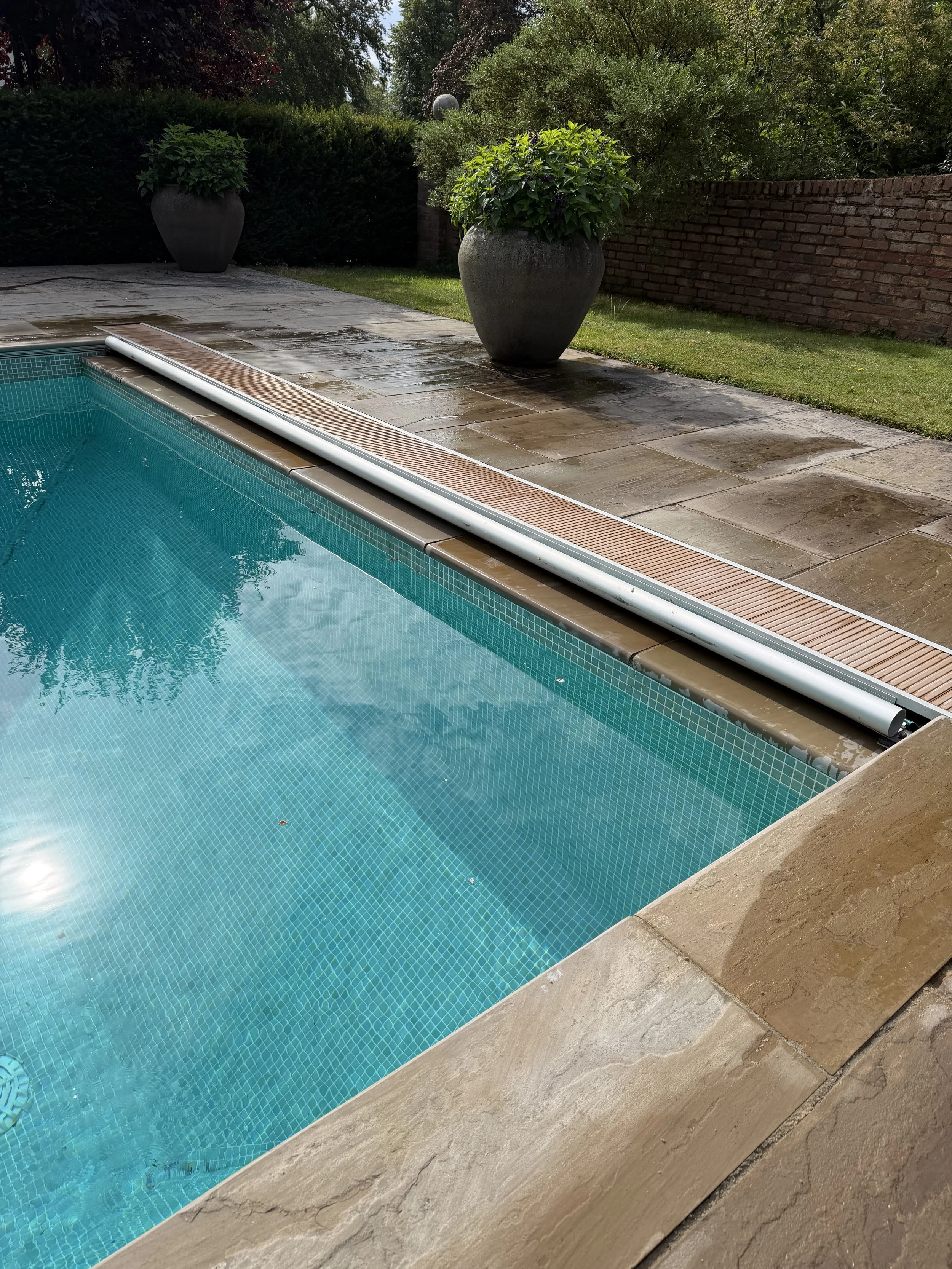 A swimming pool with light blue tiles, surrounded by a stone deck, with potted plants, a brick wall, and greenery in the background.