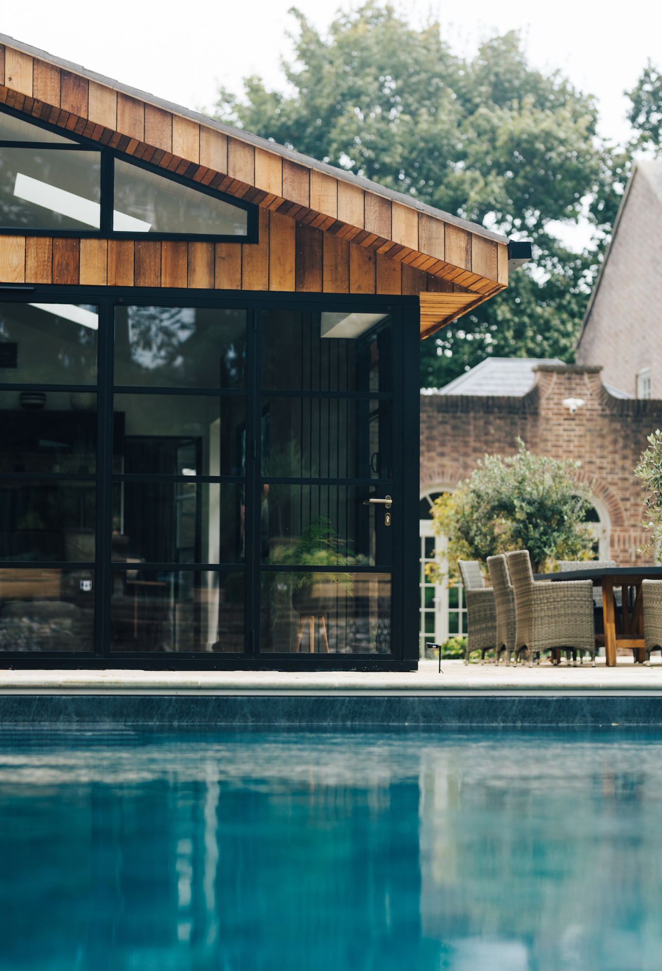 Contemporary house with large glass windows, wooden siding, outdoor patio with chairs and a table, and a swimming pool in the foreground, with trees and neighboring buildings in the background.