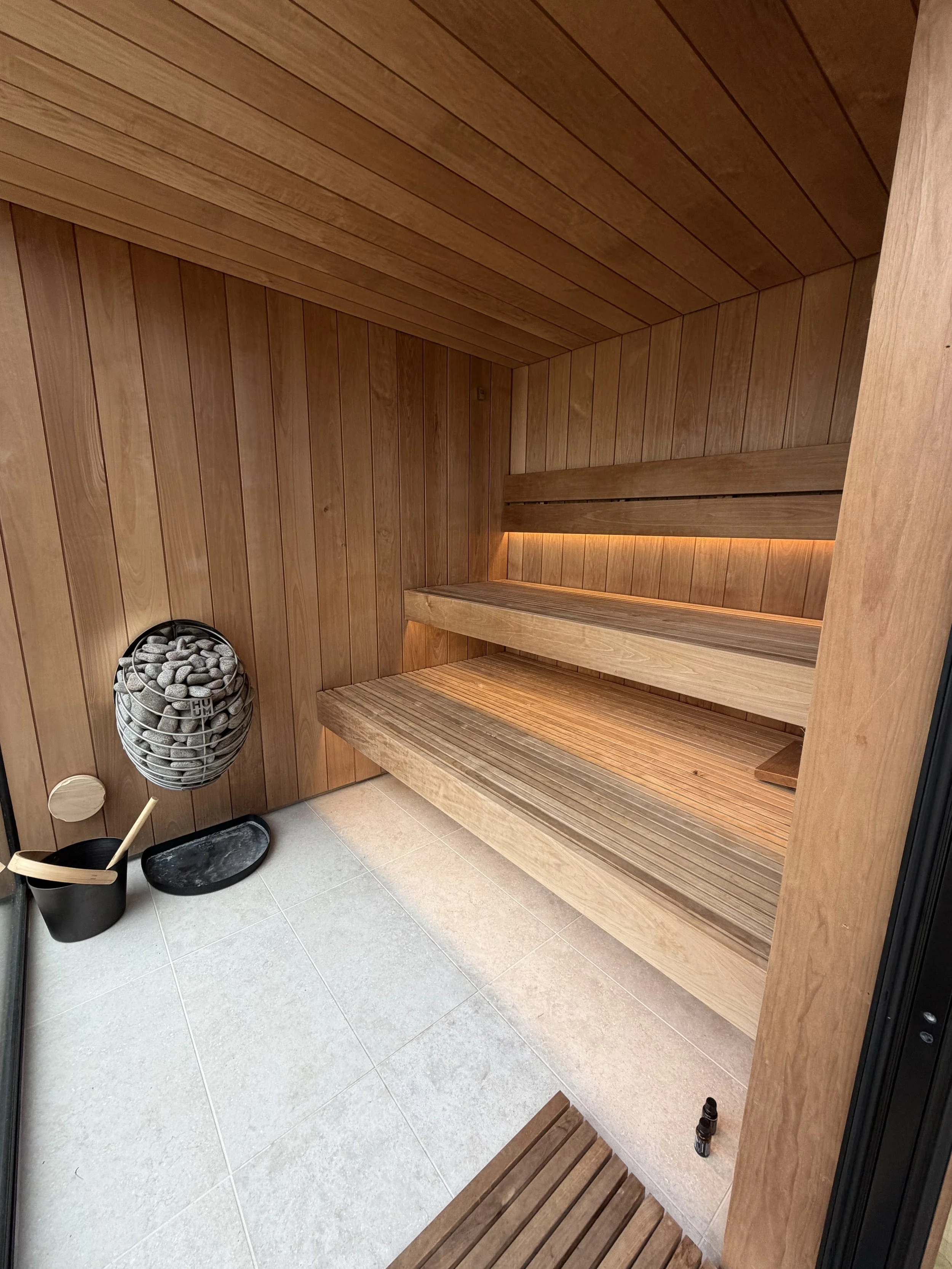 Wood-paneled sauna interior with benches, a bucket, and a ladle on the floor, and a decorative container with stones on the left side.