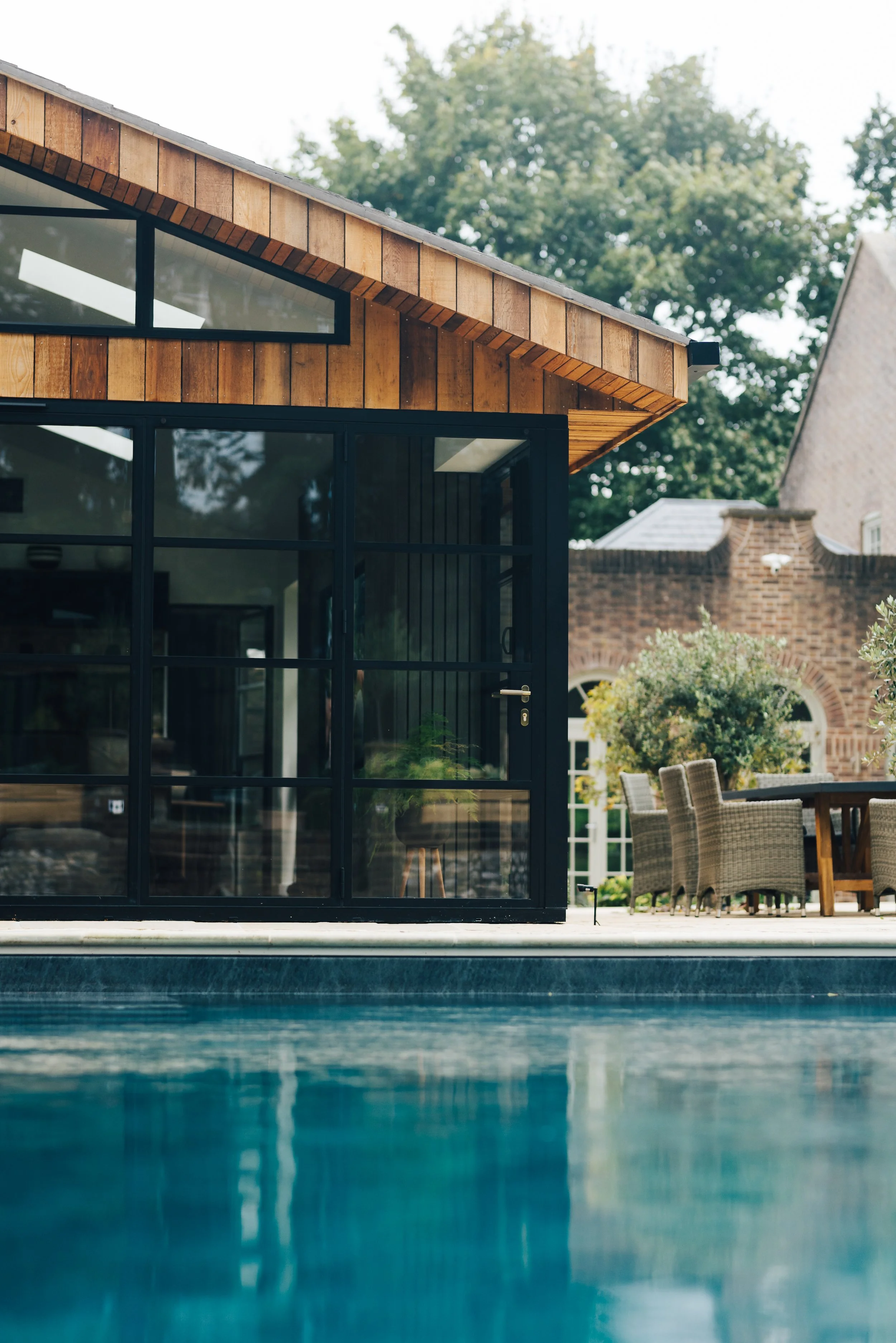 Modern house with large glass windows and wooden siding, outdoor dining area with wicker chairs, brick building in background, trees and pool in foreground.