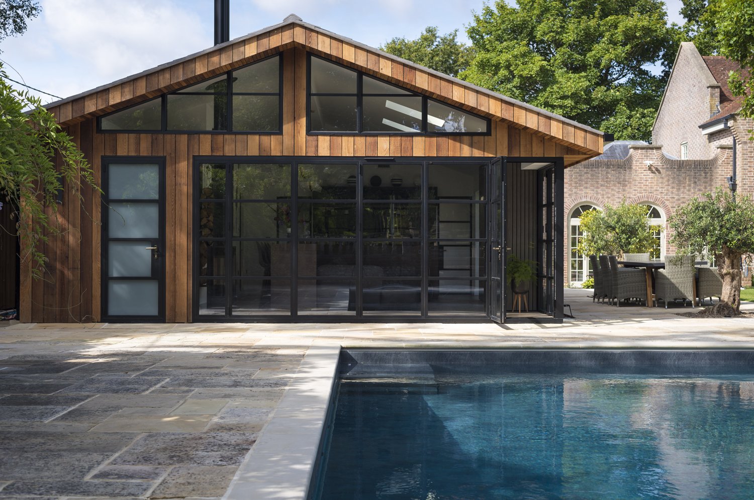 Exterior of a modern house with a swimming pool in the foreground, wooden siding, large glass windows, and a patio with outdoor furniture, surrounded by greenery and other residential buildings.