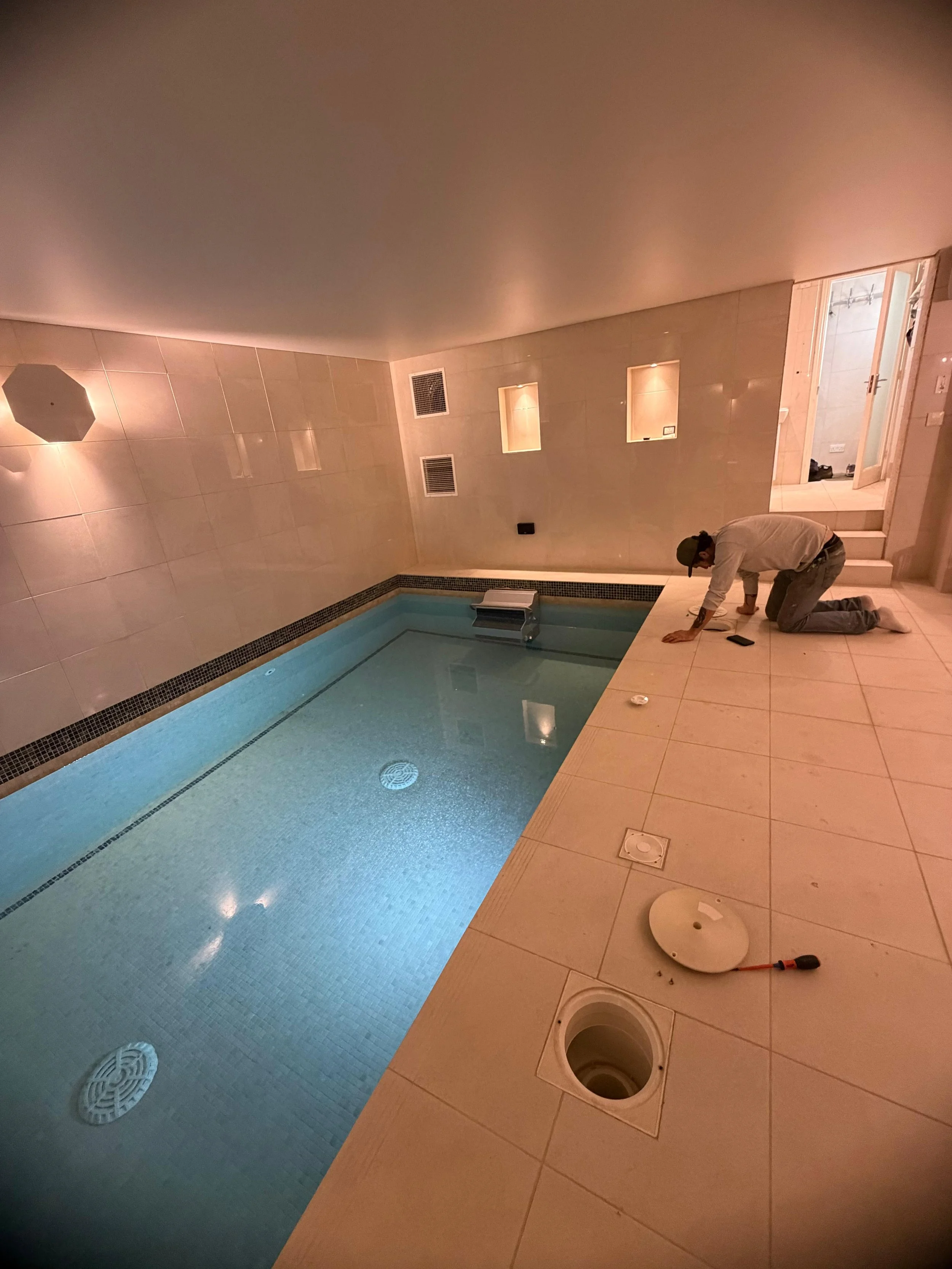 A person kneeling on the tile floor of an indoor swimming pool area, working on the installation of fixtures, with tools and plumbing parts scattered around.