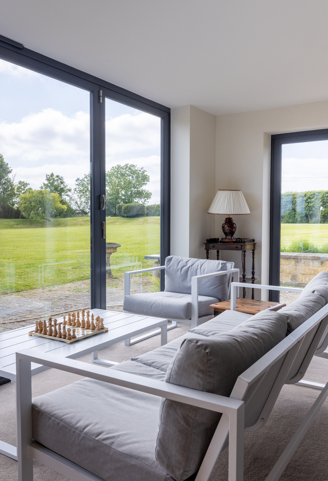 Living room with large glass sliding doors opening to a green lawn and trees outside, featuring white furniture and a chessboard on the coffee table.