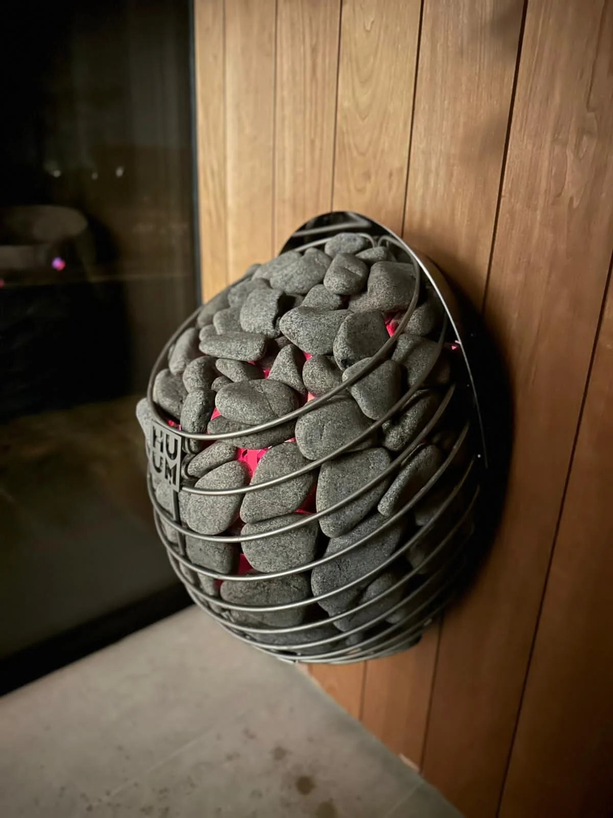 A portable electric heater filled with smooth gray stones is placed on a wooden floor near a wooden wall.