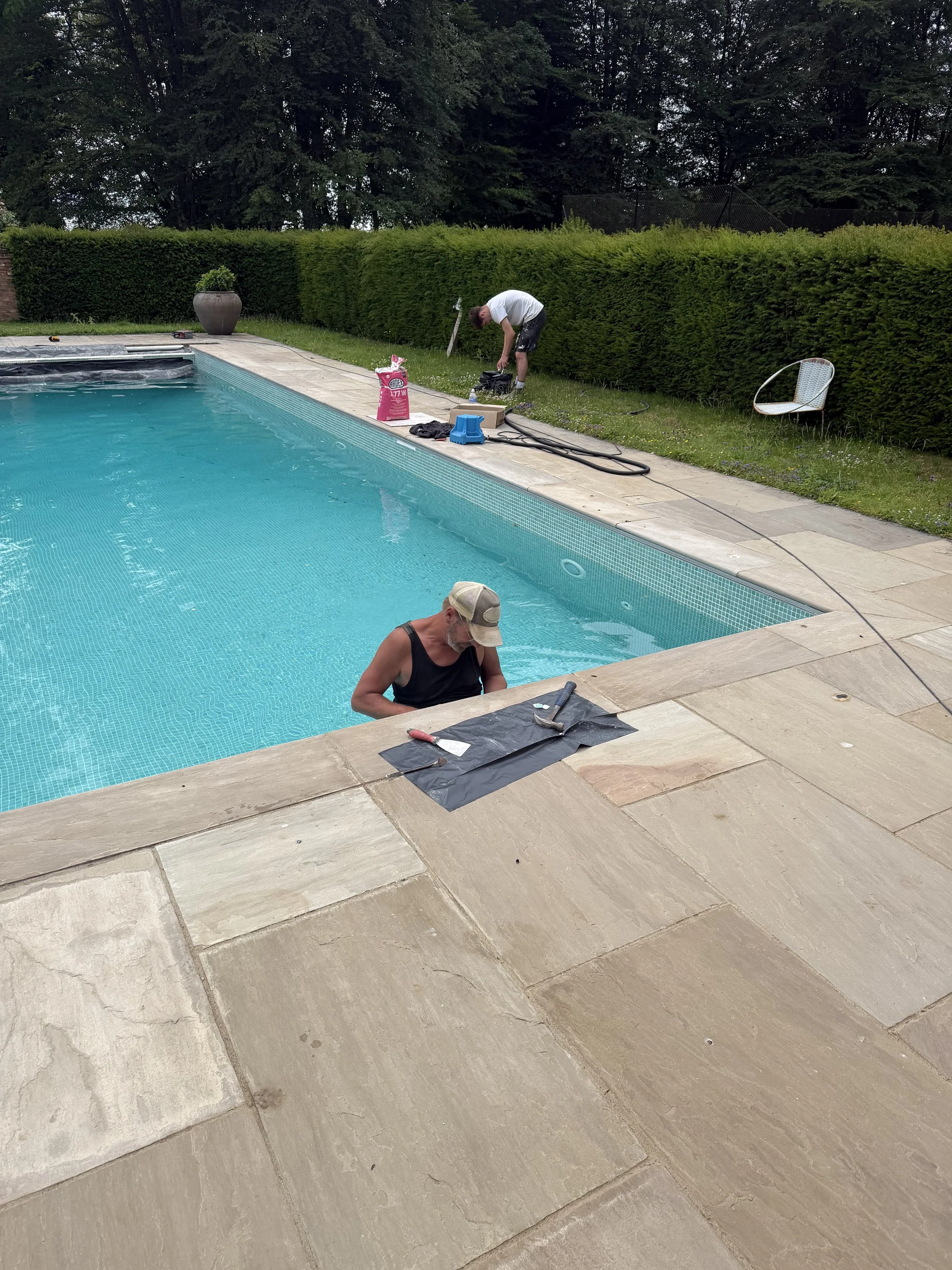 People working on a backyard pool area with tools, a water hose, and a bag of pool chemicals, surrounded by a hedge and trees.