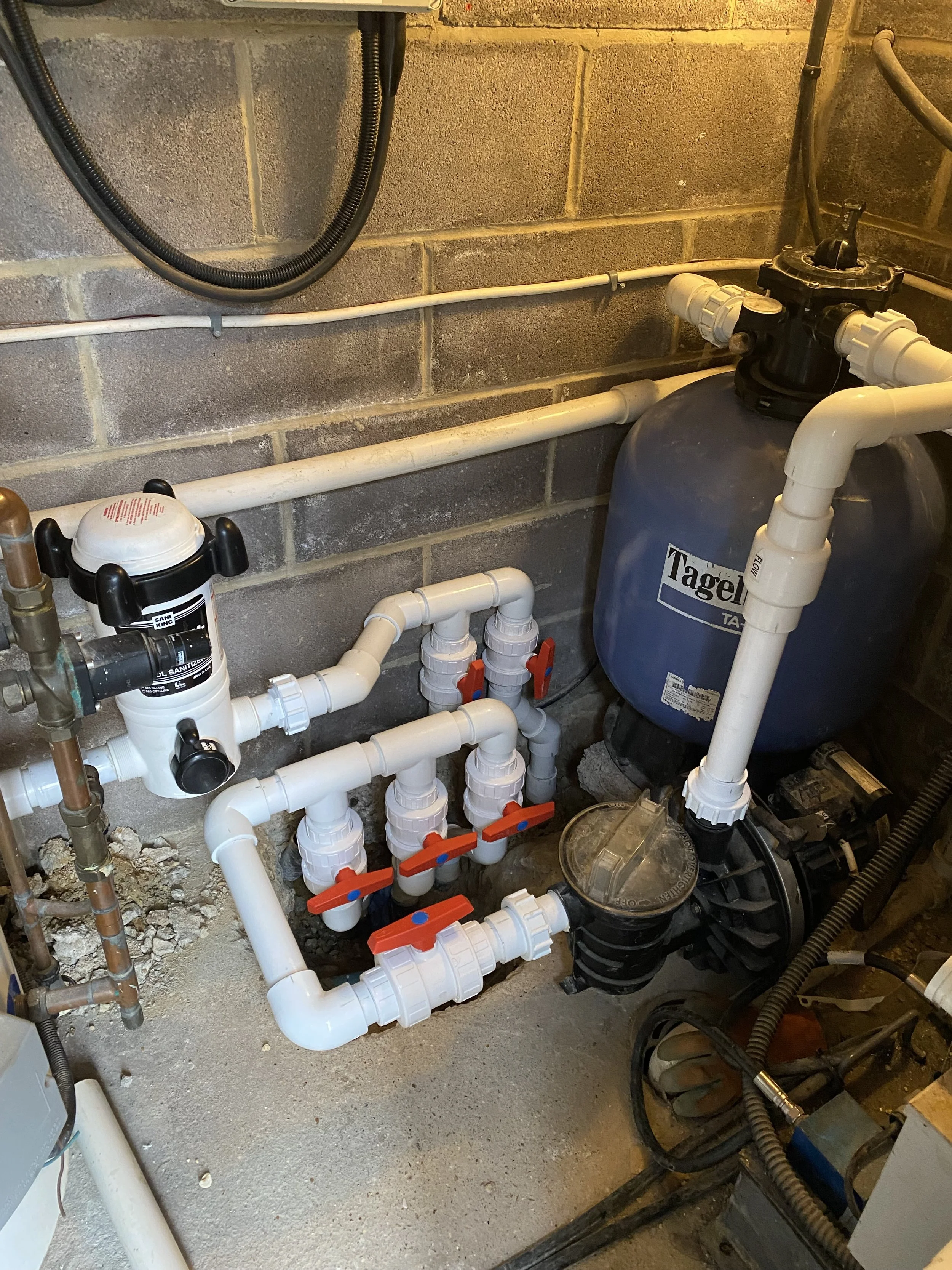 A plumbing system with white PVC pipes, valves, and a blue filter tank labeled 'Tagel'. There are several pipes connected to the filter tank and a black valve on top. The background shows a concrete block wall.