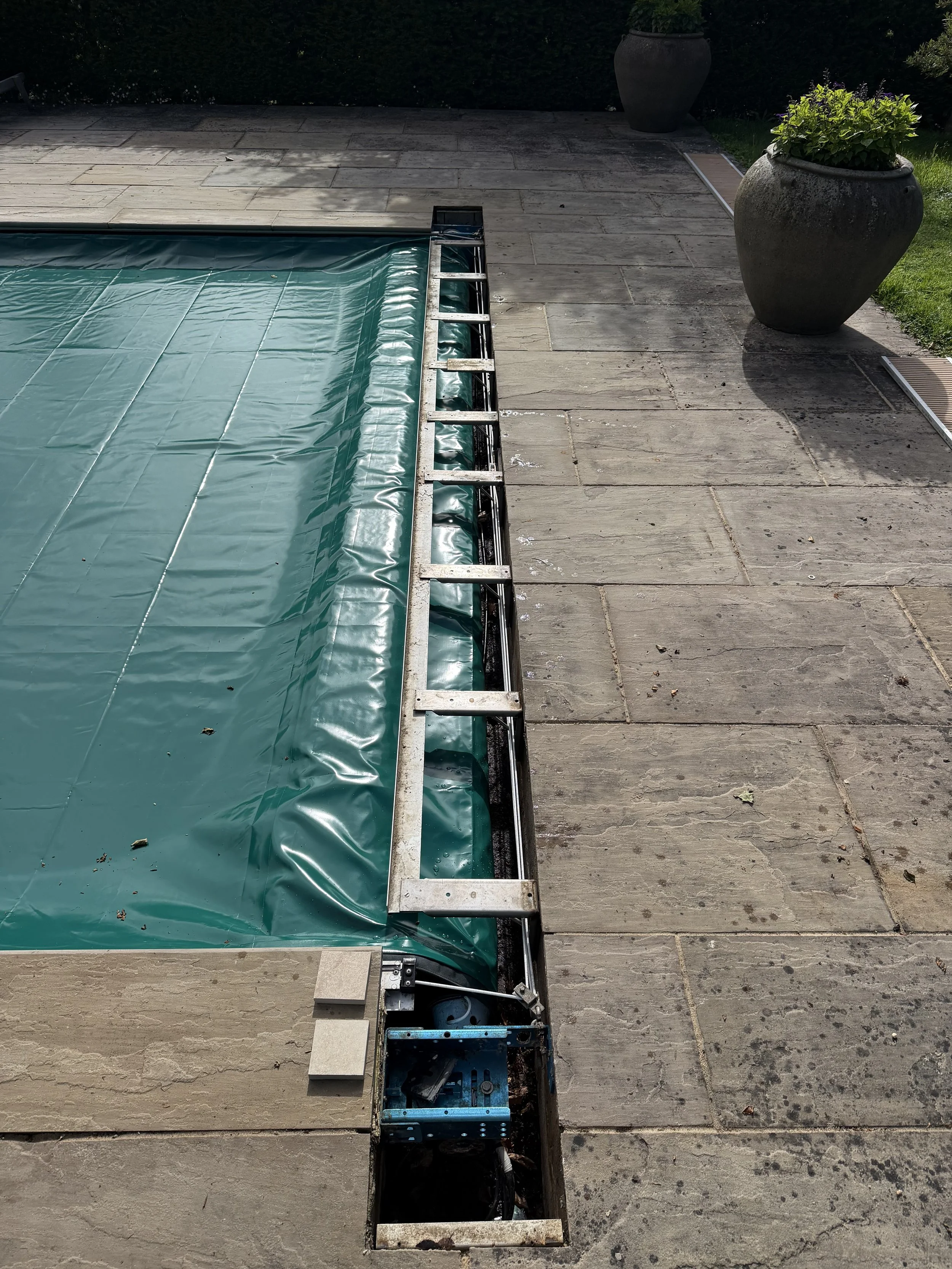 A swimming pool with a green tarp covering it, and a metal frame around the edge. The area surrounds the pool with stone tiles, and two large flower pots with plants are visible in the background.