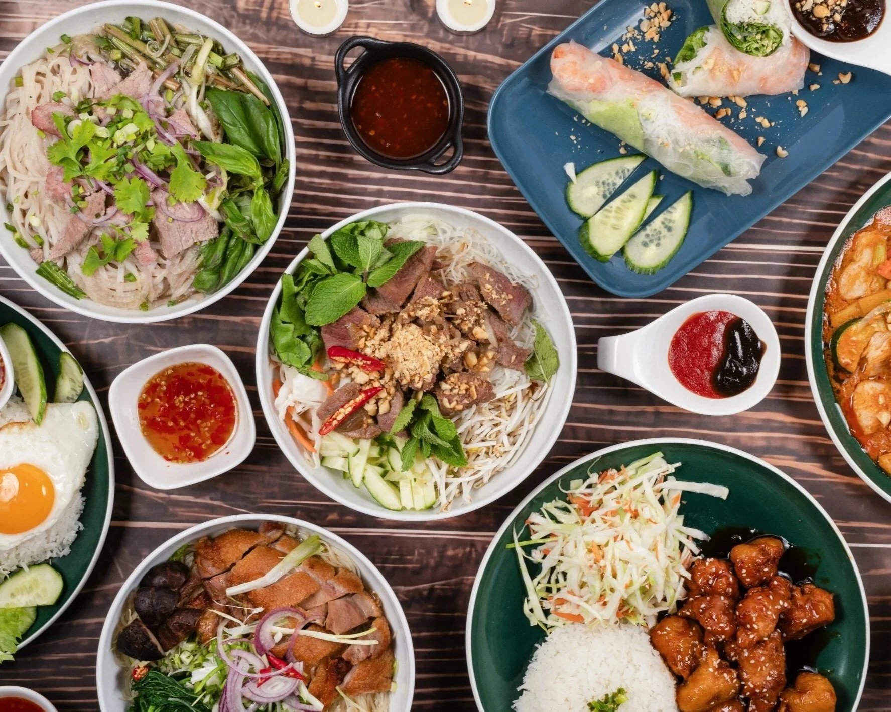 Verschiedene asiatische Gerichte auf Tellern, darunter Reis, Frühlingsrollen, Pho-Suppe, gegrilltes Rindfleisch mit Minzblättern, Gemüse, Frühlingssalat, asiatische Soßen und Süß-Sauer-Hähnchen.