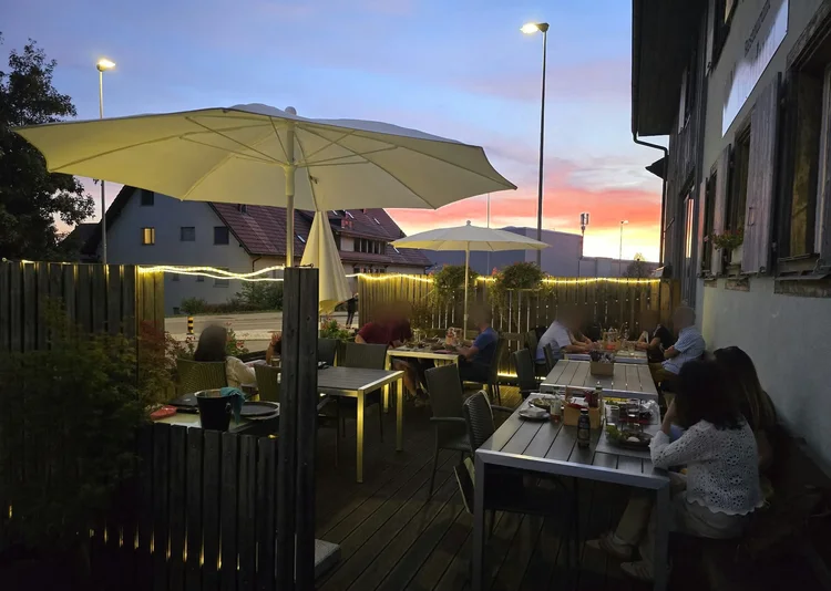 Terrasse vom Restaurant Vietdura in Tagelswangen ZH bei Sonnenuntergang: Gäste an gedeckten Tischen unter cremefarbenen Sonnenschirmen, Lichterkette am Holzzaun und rosa gefärbter Abendhimmel über den Dächern von Tagelswangen.