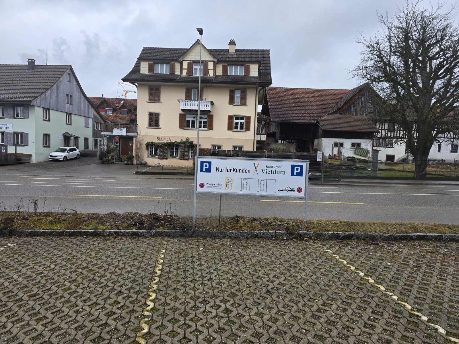 Hinweis Schild Parkplätze Restaurant Vietdura. Ansicht von hinten, Auf den Parkfeldern stehend. Im Rückgrund die Kantonsstrasse und das Restaurant VIetdura (grünes Gebäude).
