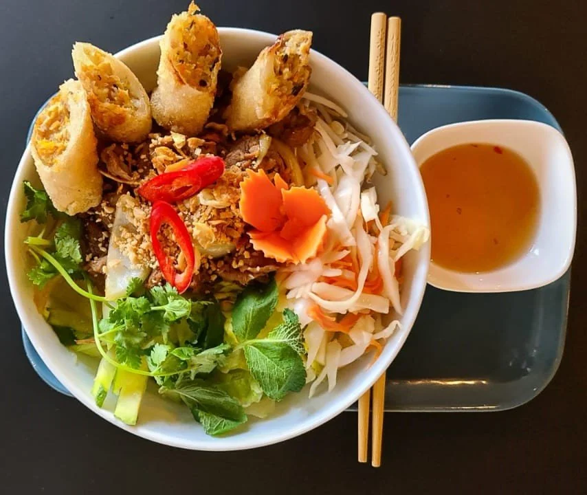Schüssel mit Pho-Nudelsuppe, Frühlingsrollen, frischen Kräutern, Chili und Gemüse, dazu ein Teller mit Brühe