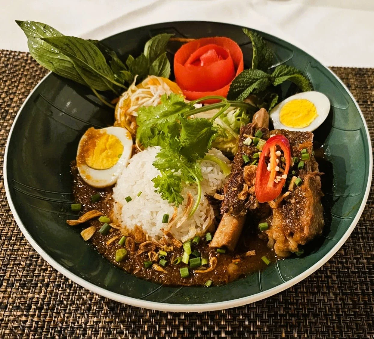 Ein Teller mit Reis, Rindfleisch, hartgekochten Eiern, frischen Kräutern und Gemüse, dekoriert mit Chili und Kartoffelrosen.