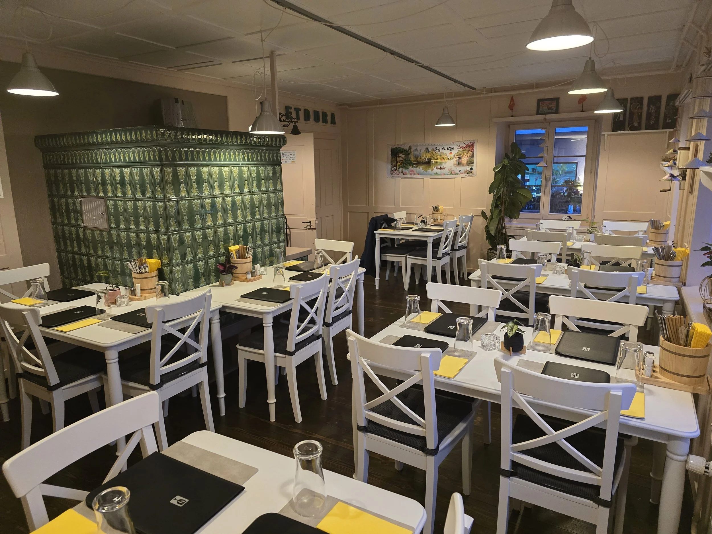 Das gemütliche Restaurant Vietdura von innen, mit weißen Tischen und Stühlen, gedeckt mit schwarzen, gelben und transparenten Gläsern, für eine Mahlzeit vorbereitet. Es gibt eine grüne Kachelofenwand und Fenster mit blauer Abendbeleuchtung.
