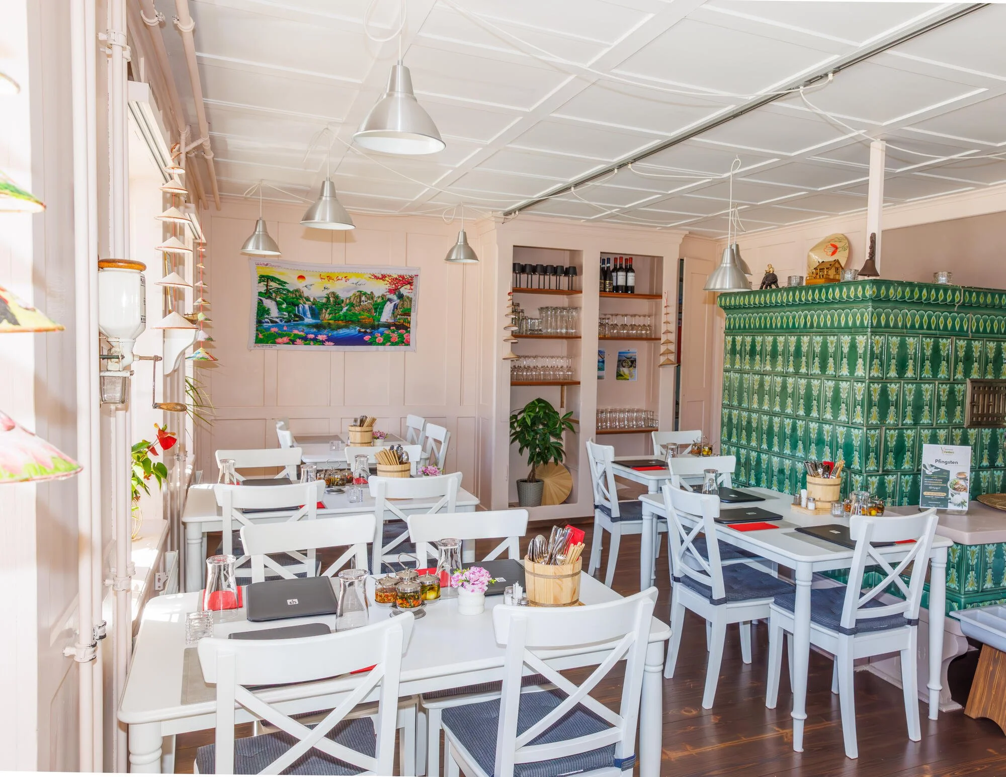 Restaurant Vietdura in Tagelswangen mit weißen Tischen und Stühlen, grüner Kachelofen, Wanddekorationen, Gläsern, Besteck und kleinen Blumenvasen auf den Tischen, mehreren hängenden Lampen und Wandregalen mit Weinflaschen und Gläsern.