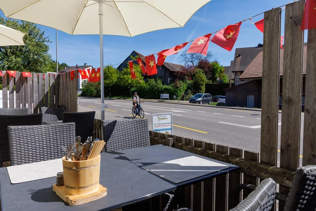 Blick auf einen Außenbereich eines Restaurants oder Cafés mit Tischen und Stühlen, Schirm und türkischen Flaggen, die an einer Schnur hängen. Im Hintergrund ist eine Straße mit einem Fahrradfahrer und parkenden Autos.