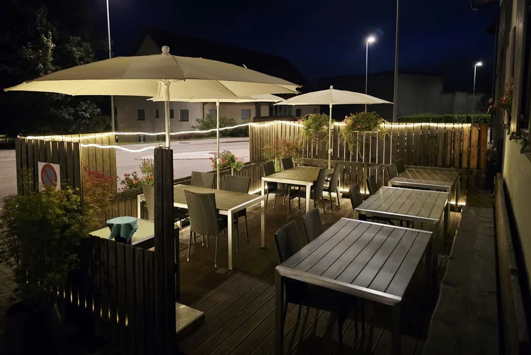 Terrasse vom Restaurant Vietdura in Tagelswangen ZH am Abend: Holzterrasse mit Tischen und Korbstühlen unter cremefarbenen Sonnenschirmen, warm beleuchteter Holzzaun mit Lichterkette und blühenden Blumenampeln.
