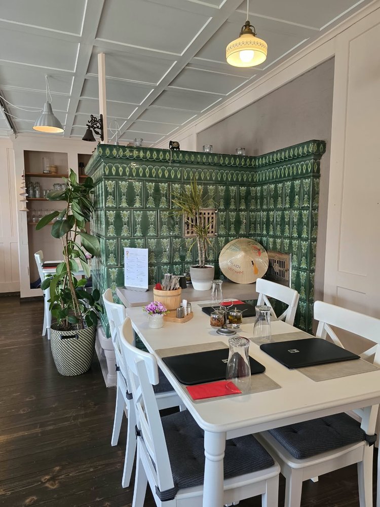 Stube vom Restaurant Vietdura in Tagelswangen ZH mit gedeckten Tischen, Zimmerpflanzen, vietnamesischen Fähnchen und dem grünen Kachelofen an der linken Wand.