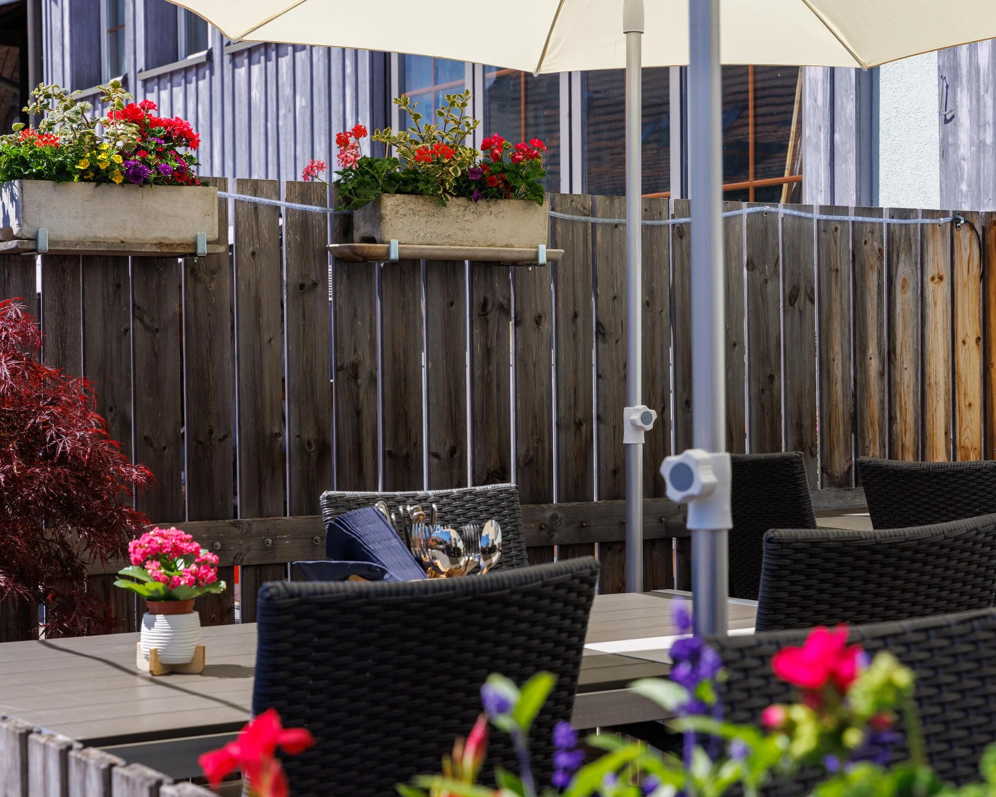 Terrasse Restaurant Vietdura in Tagelswangen mit blühenden Blumen in Pflanzkübeln, einem Sonnenschirm, einem Holzzaun, einem Tisch mit Stühlen und einem Gartenschlauch.