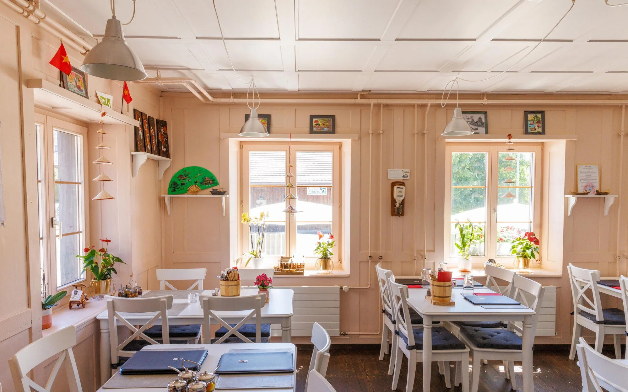 Innenaufnahme Restaurant Vietdura in Tagelswangen mit weißen Tischen und Stühlen, Pflanzen auf den Fenstern, und Dekorationen wie Vietnamesische Flaggen und Wandbilder.