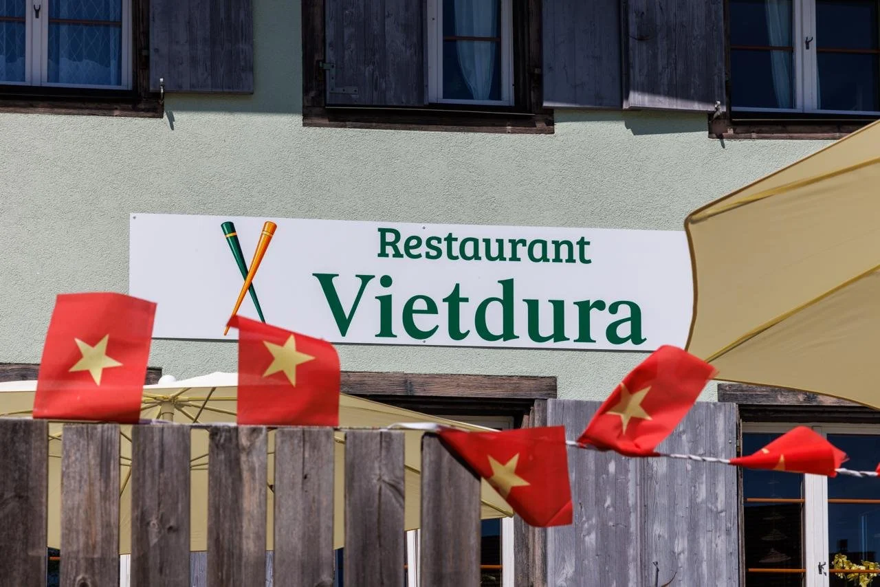 Schild mit der Aufschrift "Restaurant Vietdura" an einer grünen Hauswand, im Vordergrund rote Fähnchen mit gelben Sternen, ein Sonnenschirm.