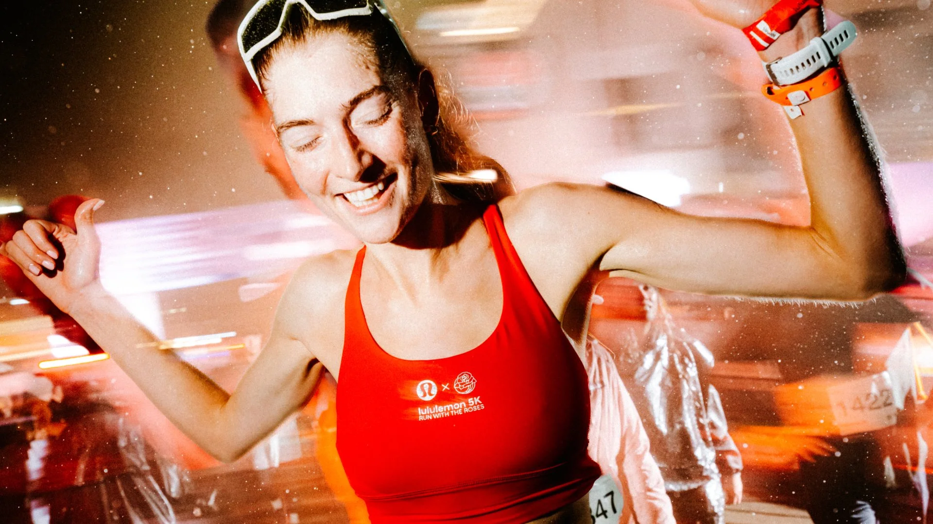 A person in a red tank top smiles as they run outside at night. A blur of lights and people are in the background.