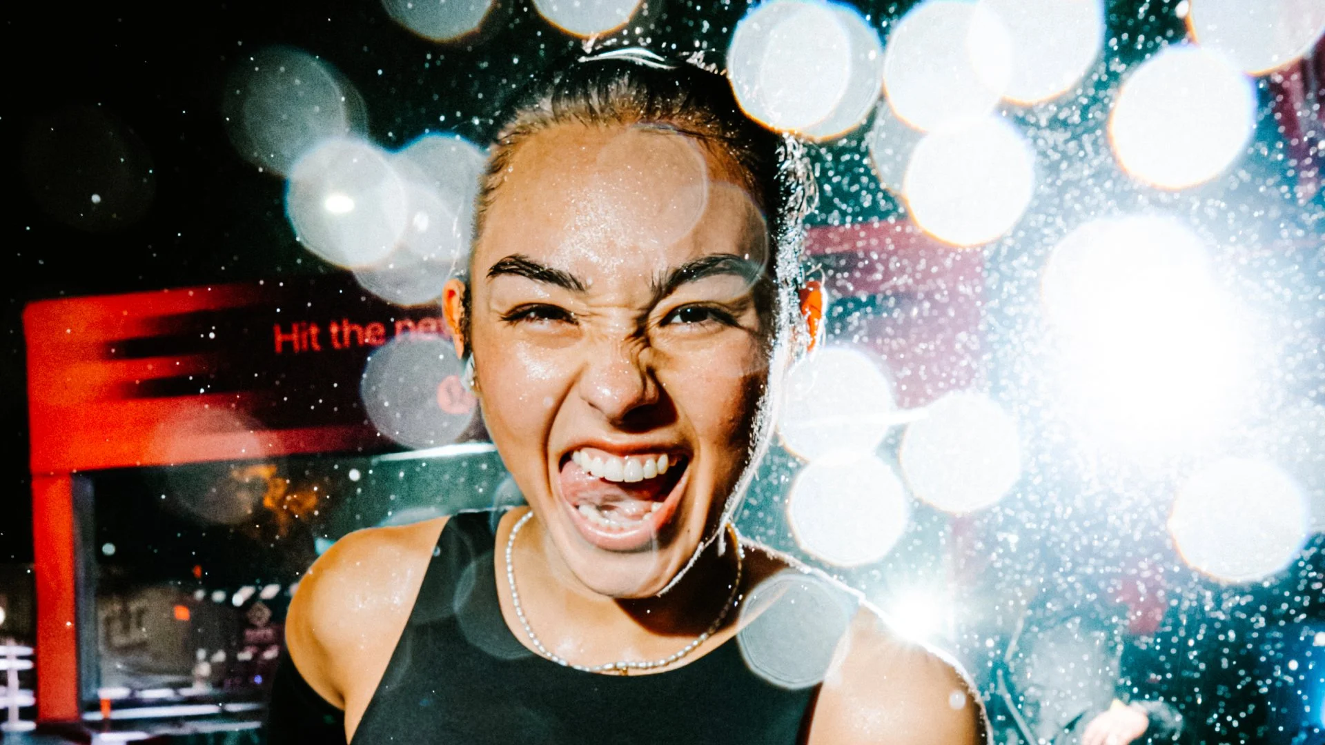 A person wearing a runner’s bib smiles with their tongue out against a backdrop of glowing lights.
