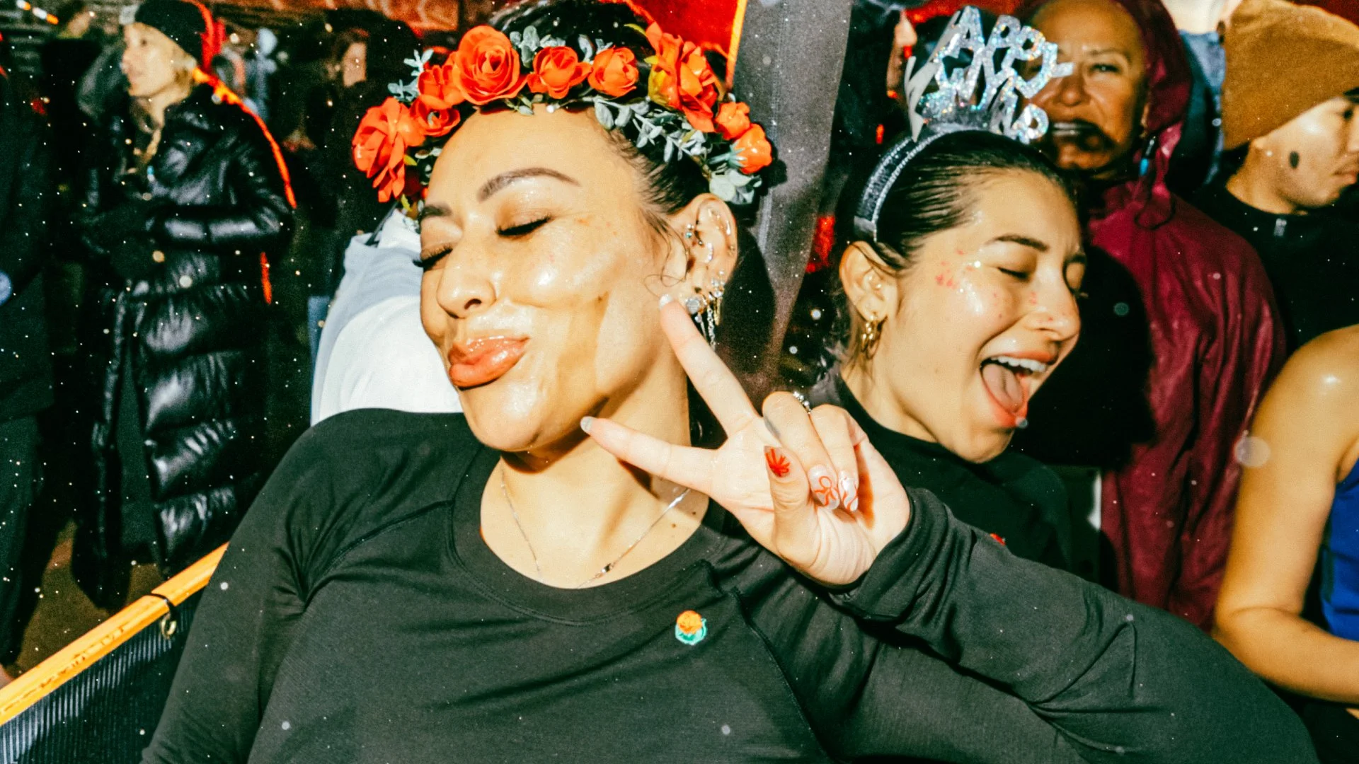 Two people smile and pose for the camera. One is wearing a flower crown and giving the peace sign, while the other wears a “happy new year” crown.
