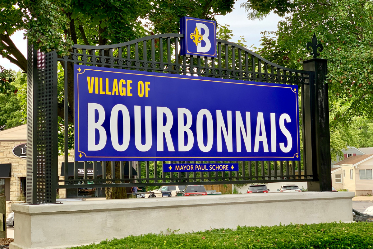 Sign for the Village of Bourbonnais with a blue background and yellow and white text, surrounded by trees and neighboring buildings.