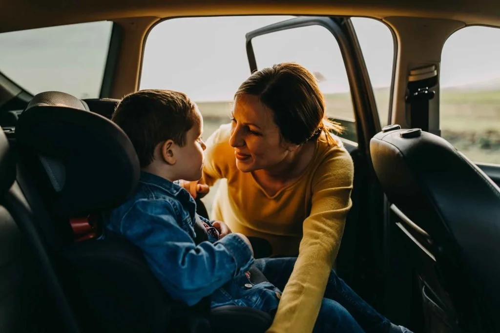 A woman and a young boy sharing a loving moment inside a car, with the woman leaning in and smiling at the boy who is seated in a car seat.