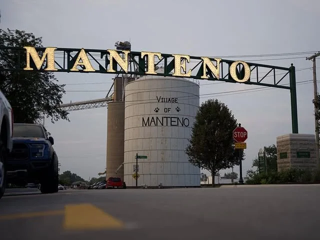 Entrance sign for the village of Manteno with large letters crossing over the road and a large silo in the background.