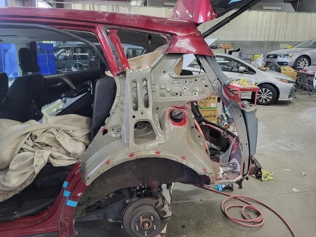 A partially disassembled red SUV in an auto repair shop, showing exposed metal framework and missing rear quarter panel. The interior is visible with seats and a tarp. Other cars and repair equipment are present in the background.