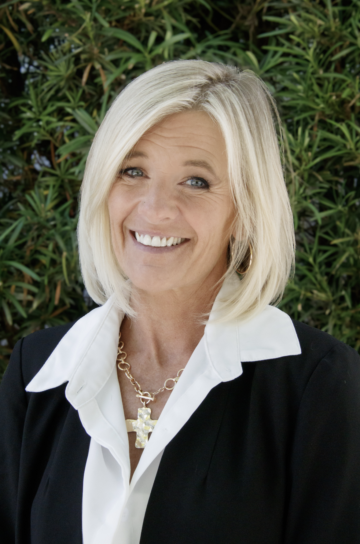 A smiling middle-aged woman with blonde hair and blue eyes, wearing a white collared shirt, a black blazer, a gold necklace with a star pendant, and hoop earrings, standing outdoors in front of green leafy bushes.