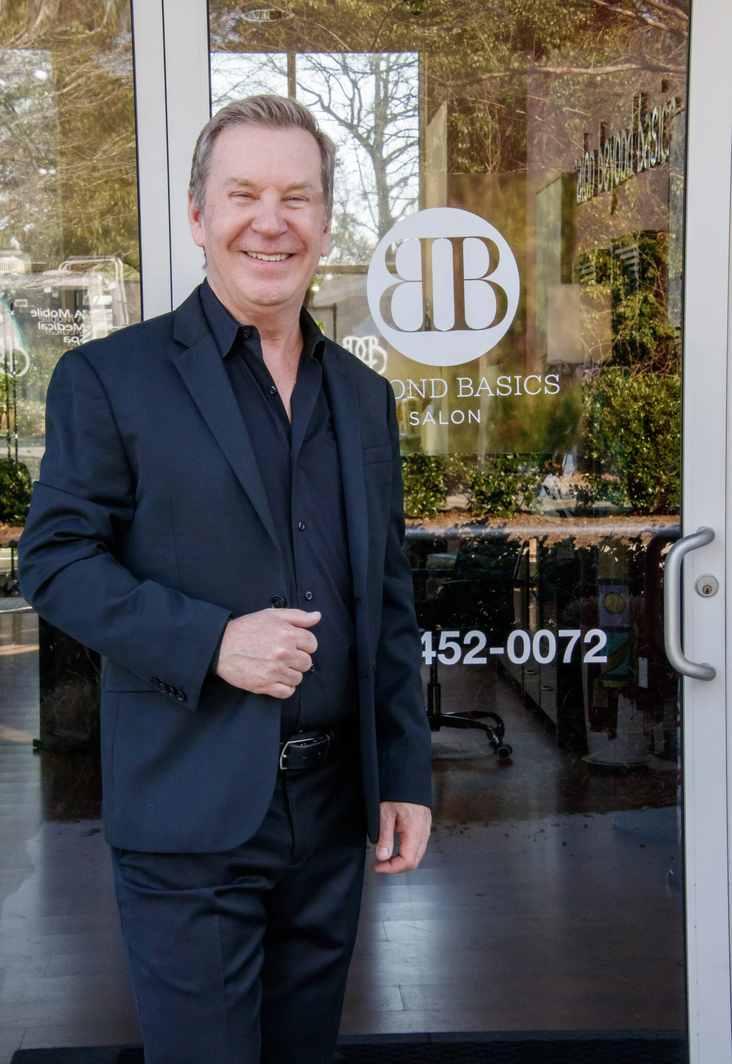 Smiling man in a black suit standing outside salon entrance with glass door featuring 'Second Basics Salon' logo, trees reflected in window.