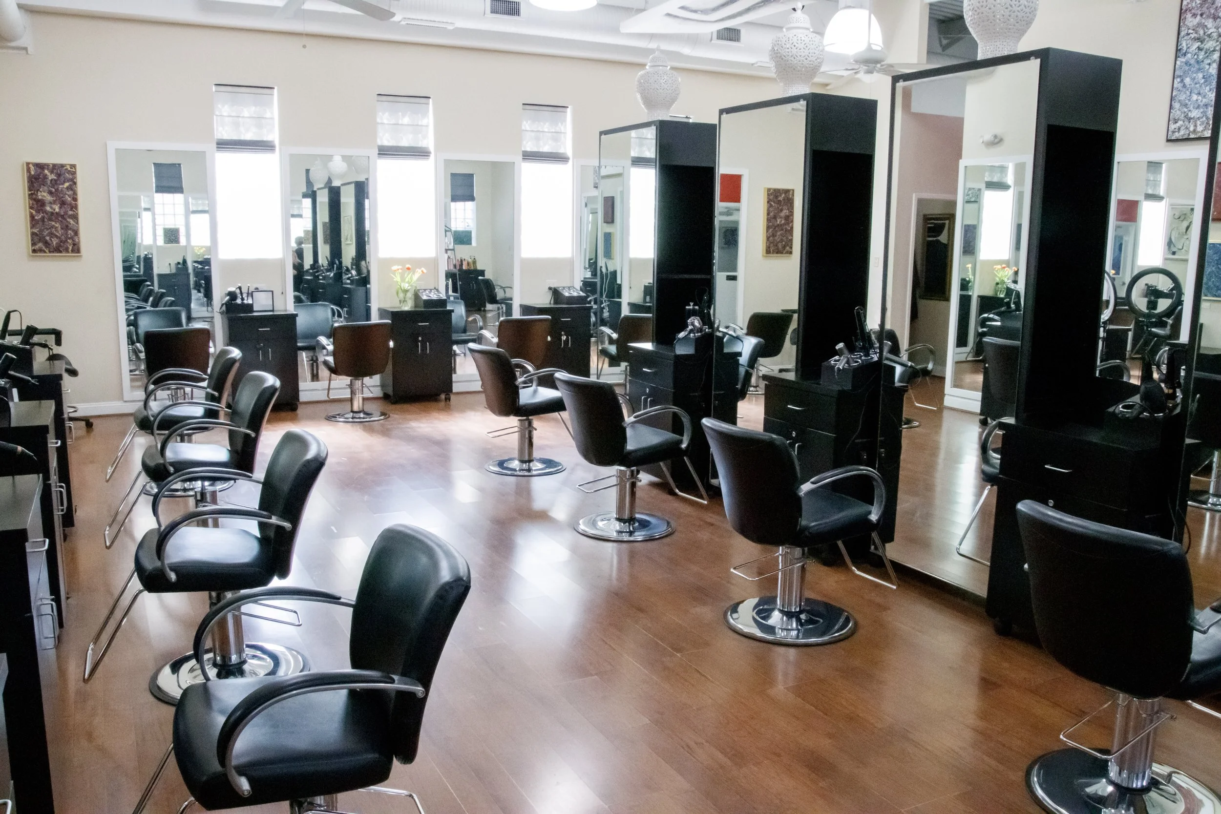 Empty hair salon with black styling chairs, mirrored stations, and large windows letting in natural light.