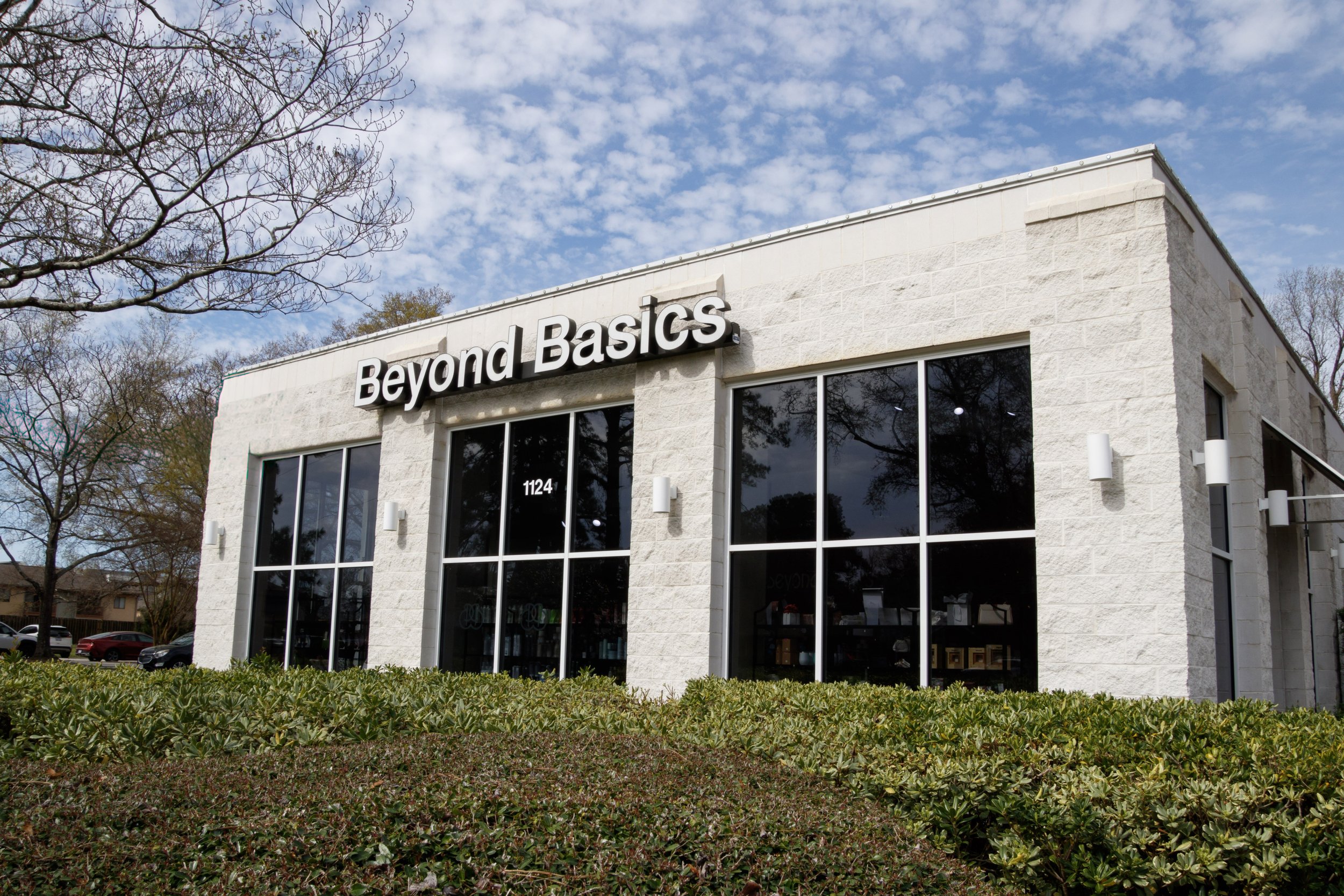 Exterior view of the Beyond Basics building with large glass windows, white stone facade, and a sign with the name at the top, surrounded by neatly trimmed bushes and trees with bare branches under a partly cloudy sky.