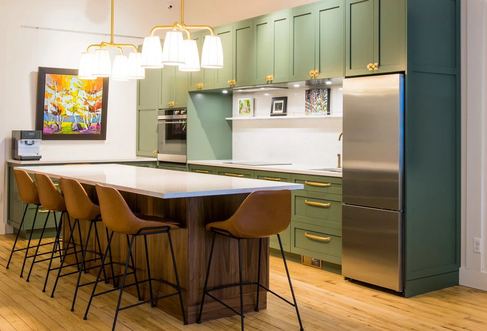 A modern kitchen with green cabinets, a wooden island and pendant lights. A colorful painting from Colborne Street Gallery hangs on the wall. Cream paint on the walls is from Farrow and Ball.