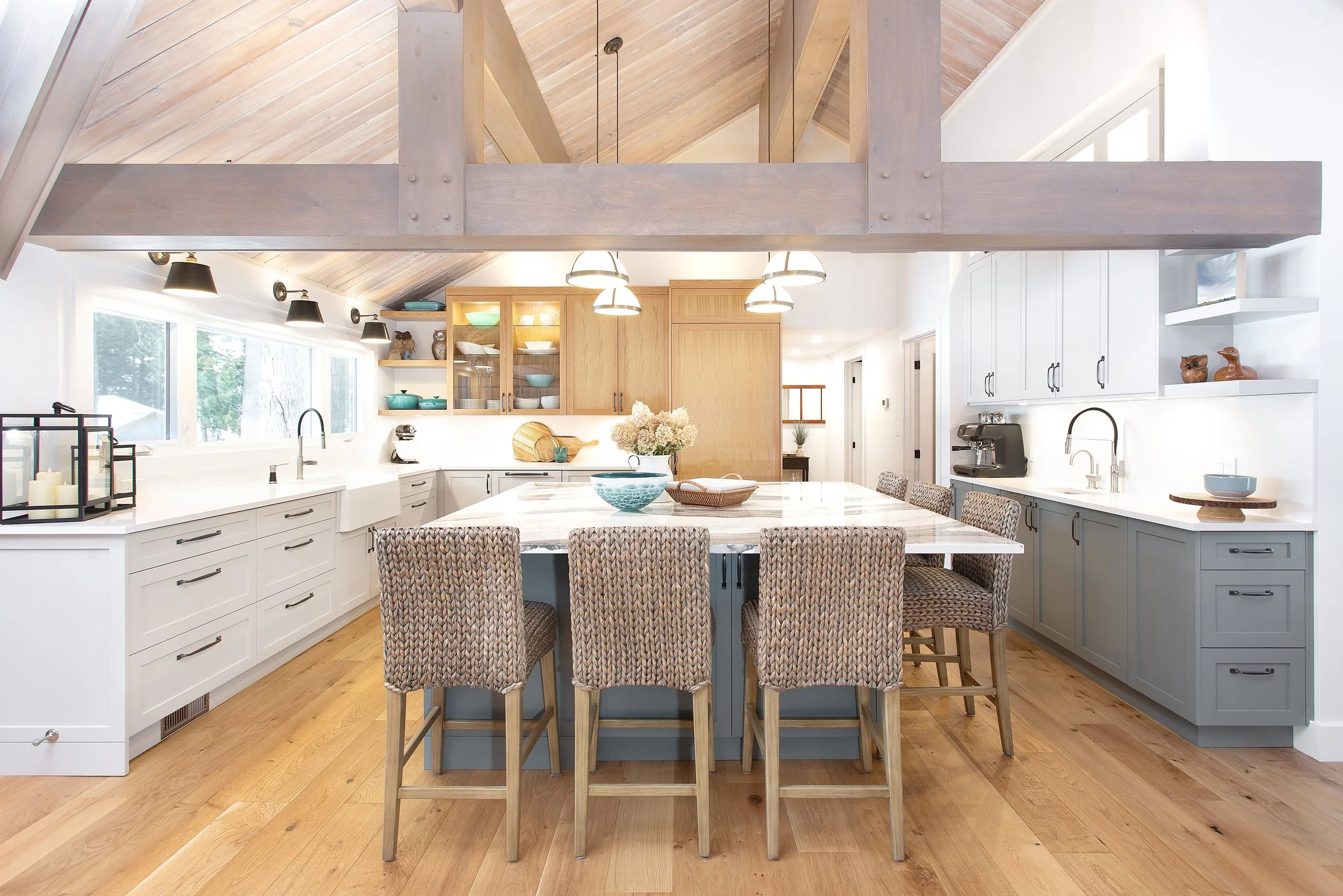 A custom designed bright and spacious kitchen by Lucvaa Kitchens and Home by Tim and Chris. White and blue-gray cabinetry and Myoh cabinet handles. Modern pendant lighting hangs over a large island with a white countertop by Cambria and bar stools.