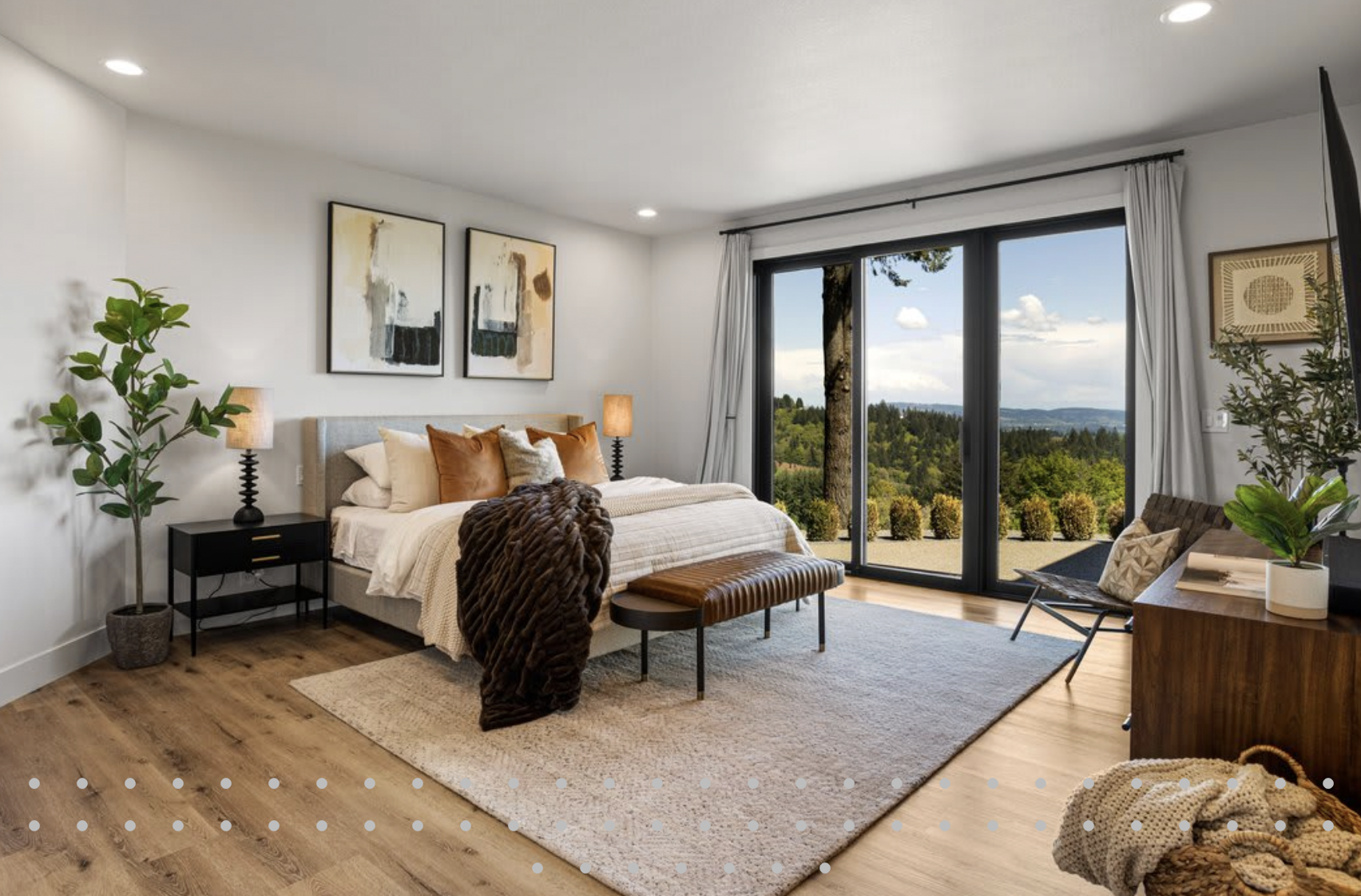 Modern bedroom with large sliding glass doors overlooking a green landscape, featuring a bed with beige and brown pillows, wooden furniture, indoor plants, and abstract wall art.