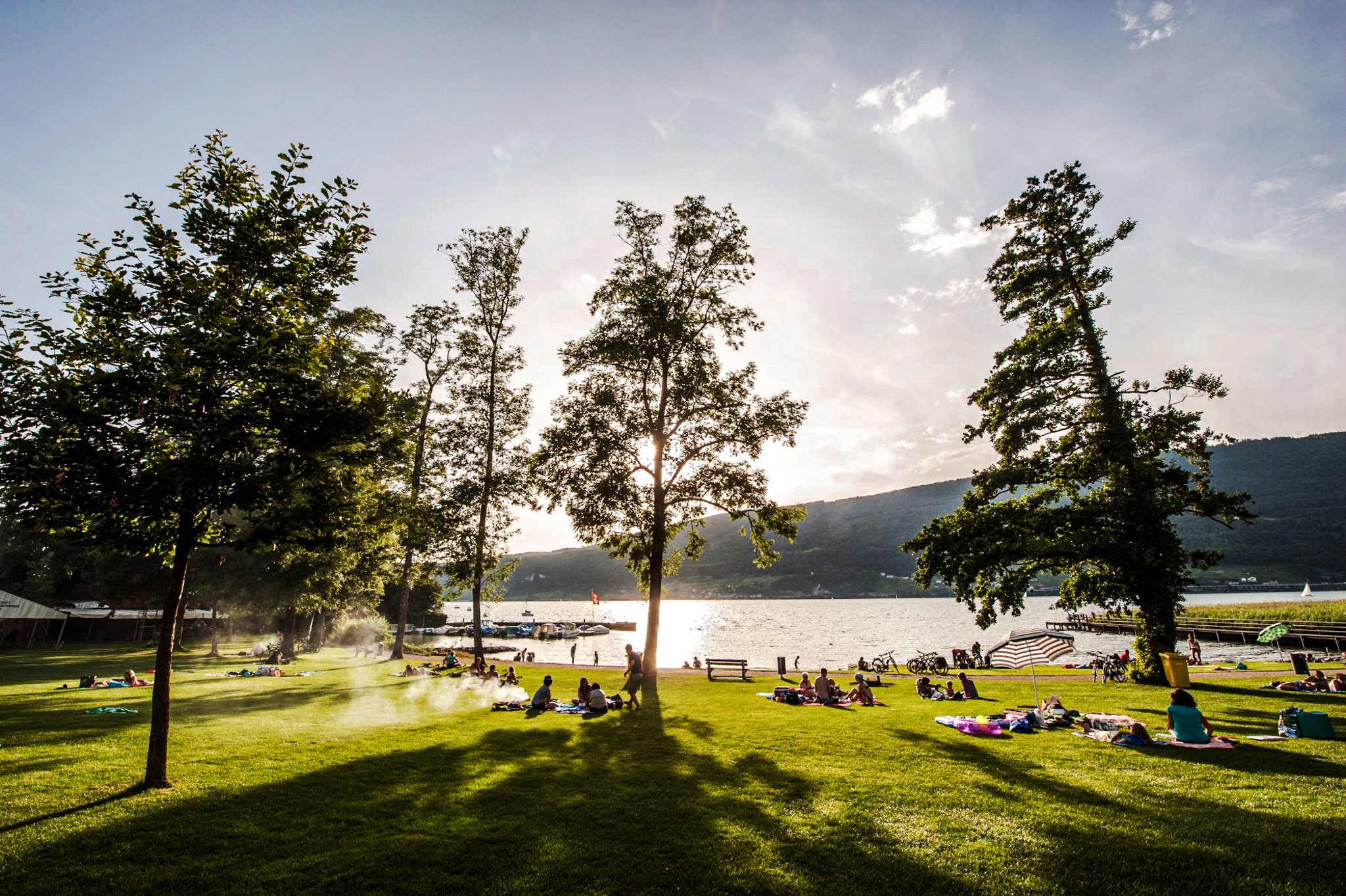 Menschen entspannen im Park am Seeufer, Liegen auf der Wiese, Bäume bieten Schatten, Hintergrund Berge und Wasser, sonniger Tag.