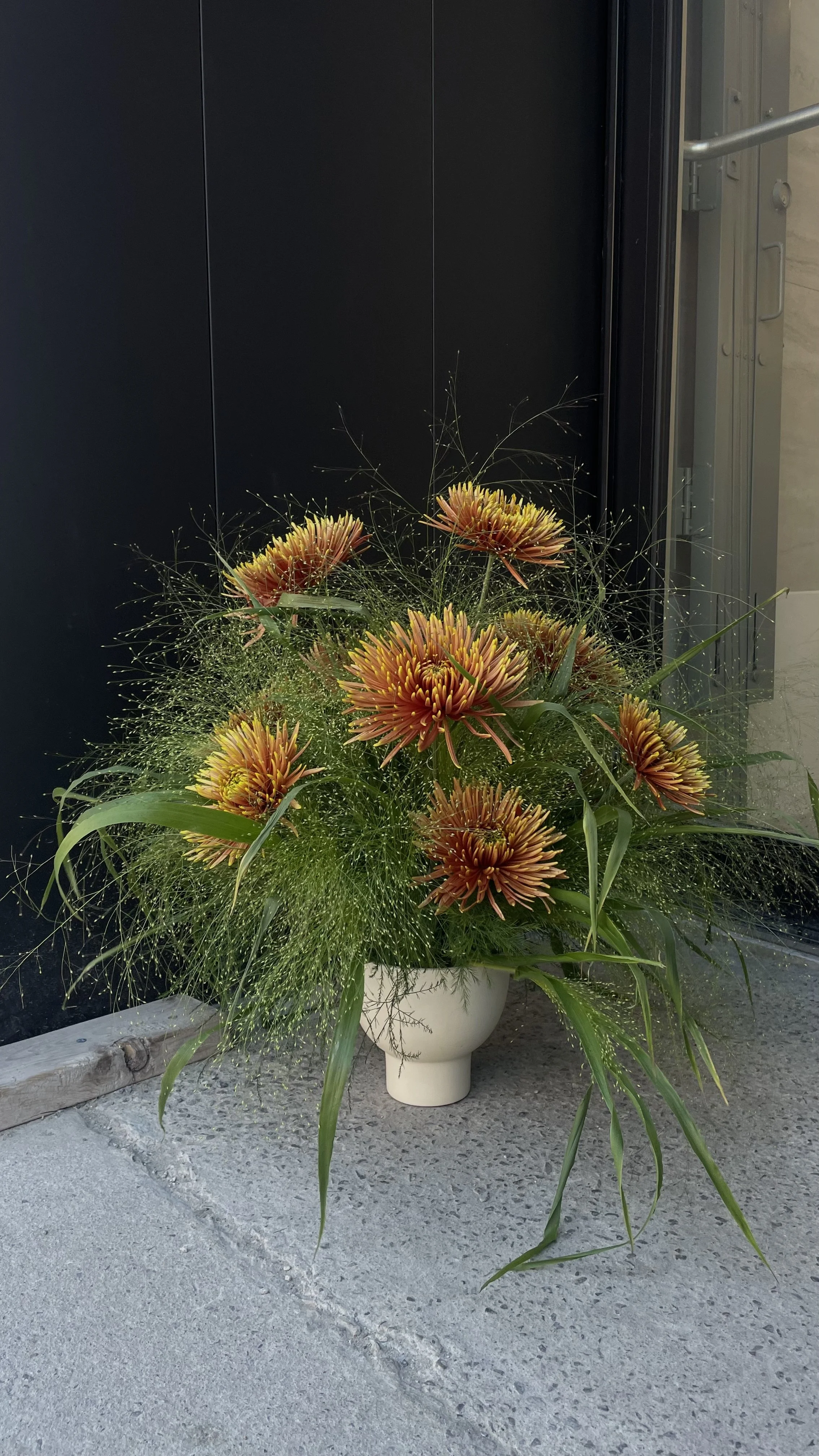 Un arrangement floral avec des grandes fleurs orange et jaune dans un pot blanc, placé sur un sol en béton, devant une porte noire.