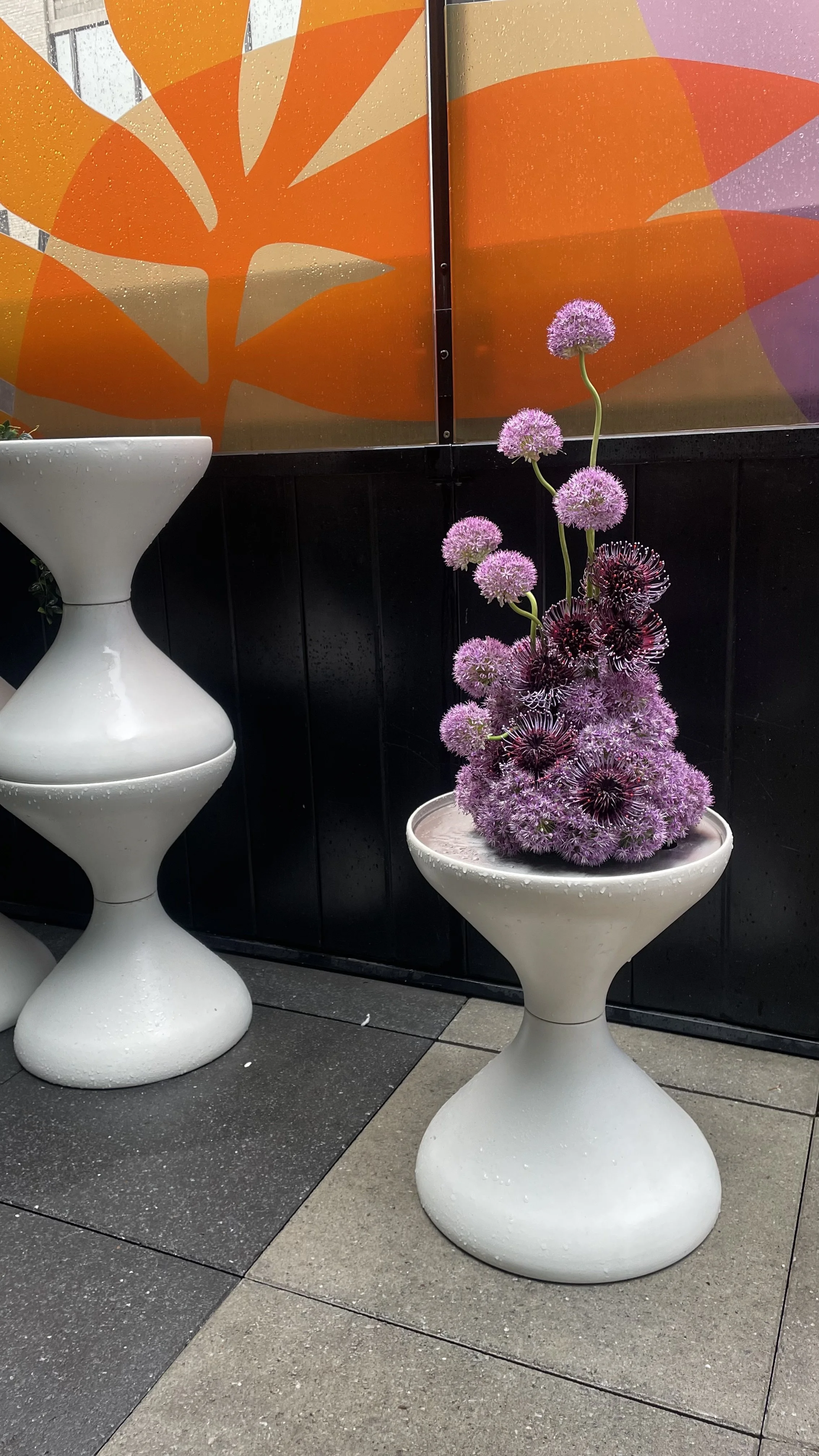 Vase avec des fleurs violettes en forme de globes, placé sur une table blanche. En arrière-plan, un mur avec un panneau en verre coloré orange et rose.