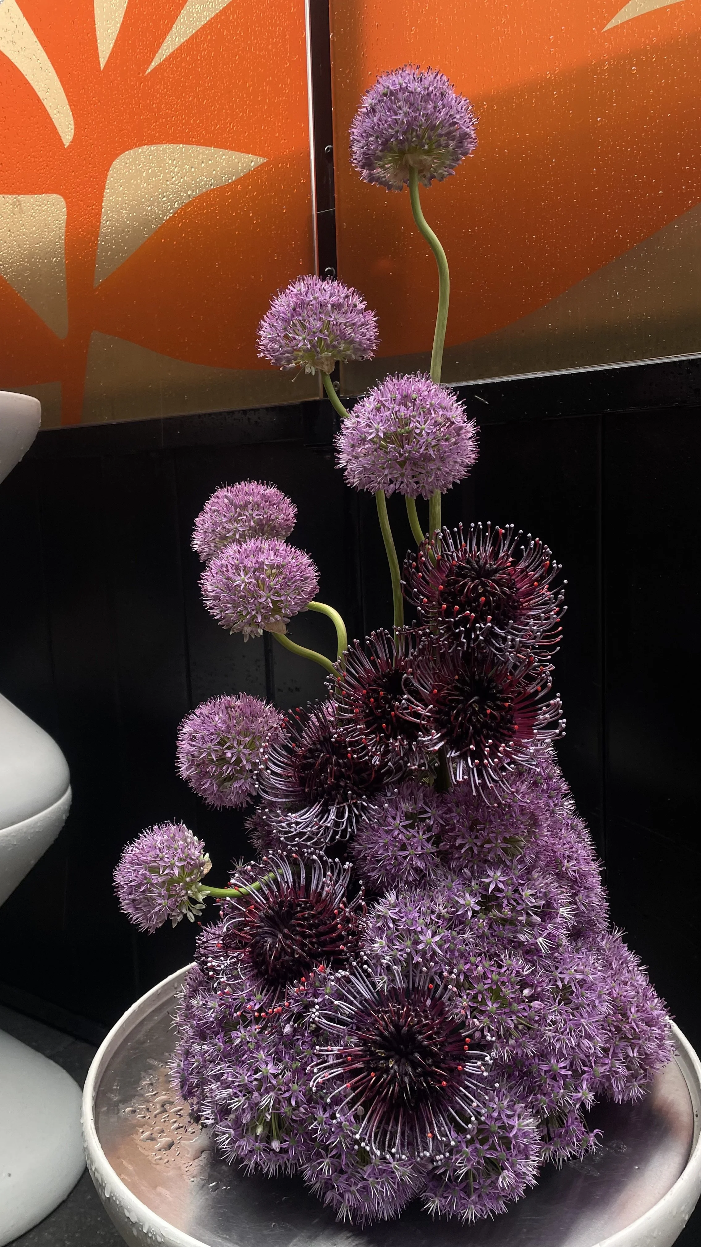 Un bouquet de fleurs violettes et noires dans un grand vase en métal, avec un fond orange et noir.