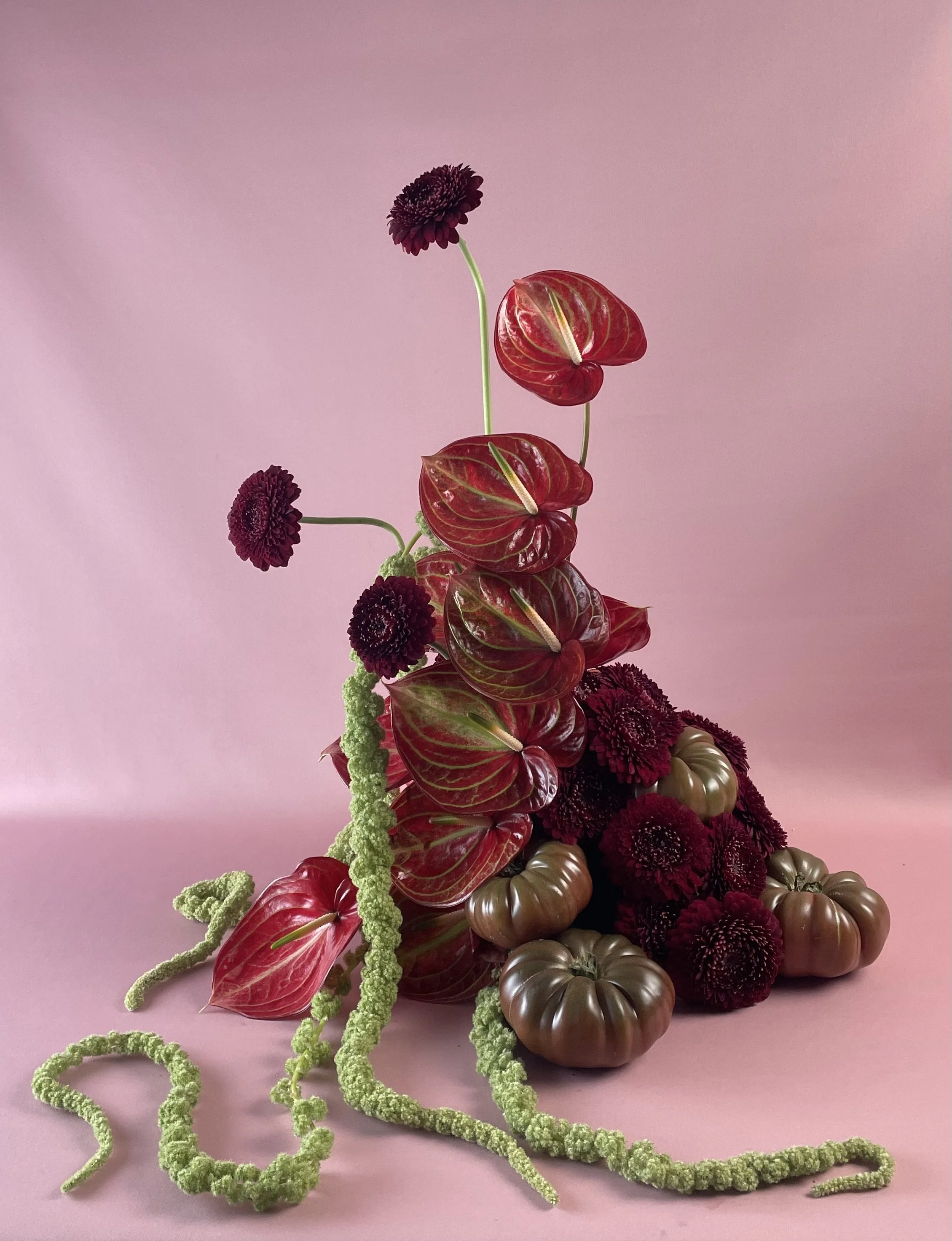 Composition florale avec des fleurs rouges et verts, des tomates, et des plantes séchées sur un fond rose clair.