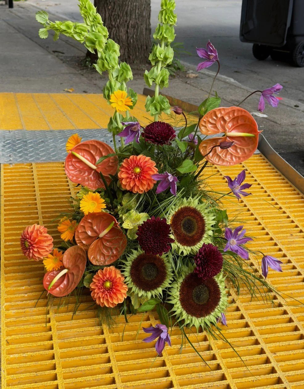 Un arrangement floral coloré avec des callas roses, des tournesols, des dahlias, des zinnias, des iris violets et d’autres fleurs, placé sur une grille jaune dans un environnement urbain.