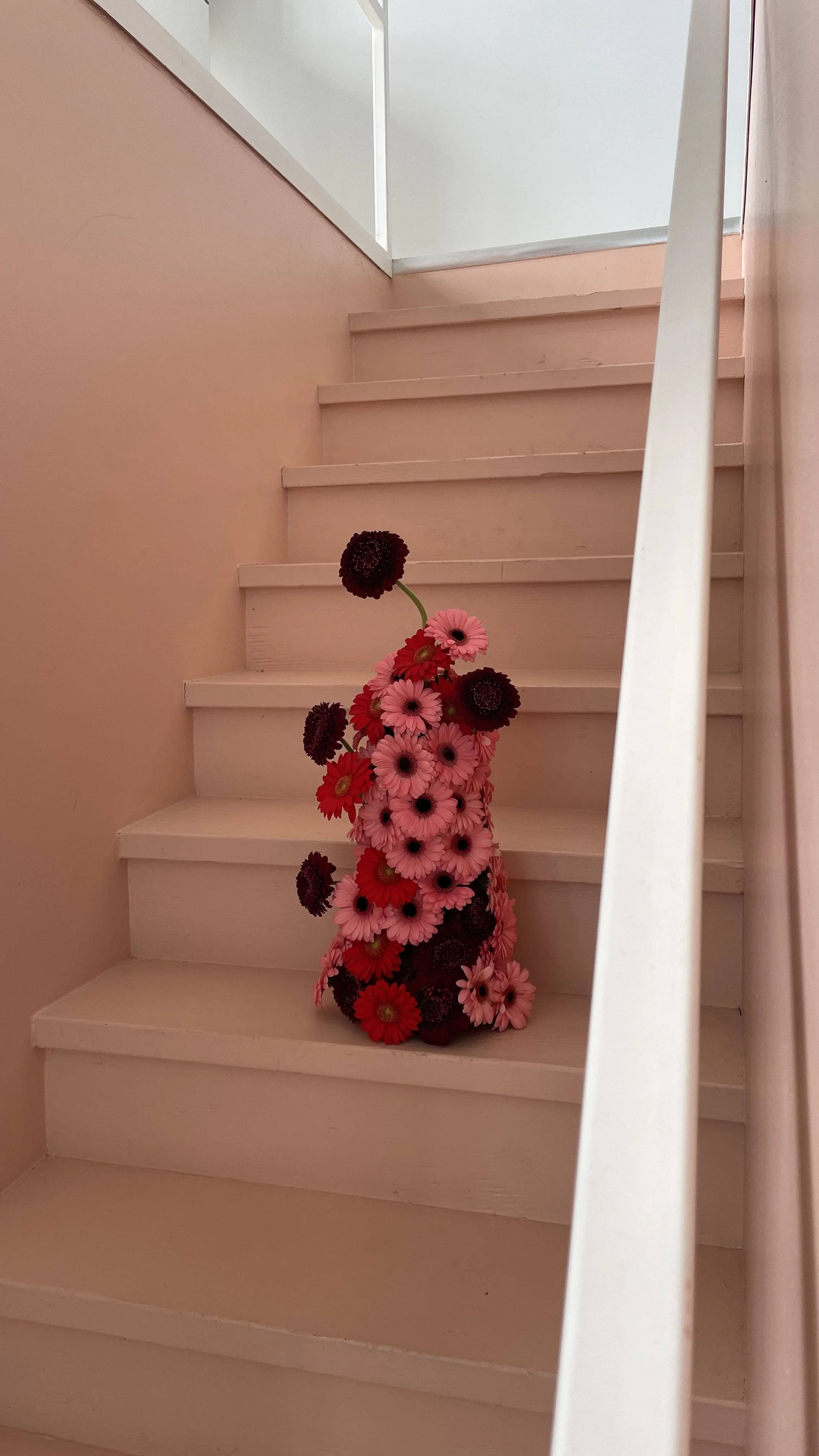Un vase rempli de fleurs roses, rouges et sombres placé sur un escalier en bois peint en blanc.