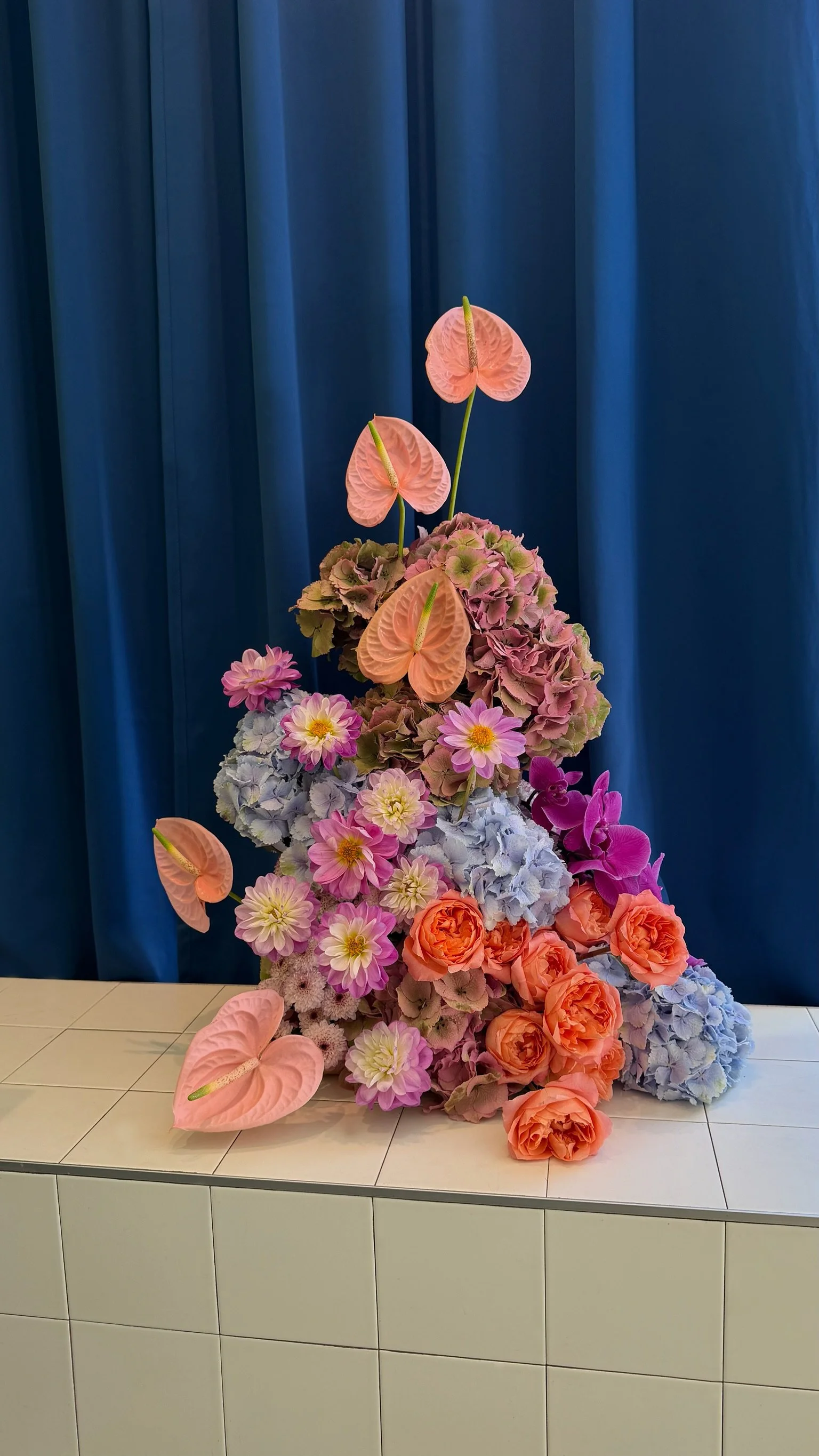 Arrangement floral composé de différentes fleurs roses, orange et violet avec des anthuriums roses, des hortensias, des dahlias et des roses, placé sur une surface en carrelage blanc devant un rideau bleu.