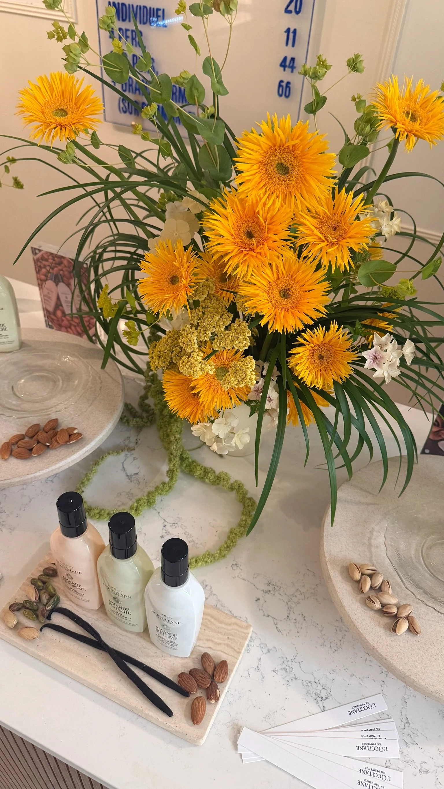 Un arrangement floral avec des fleurs jaunes et blanches, des feuilles vertes, placé sur une table blanche avec des produits de soin, des pistaches et des papiers d'information.