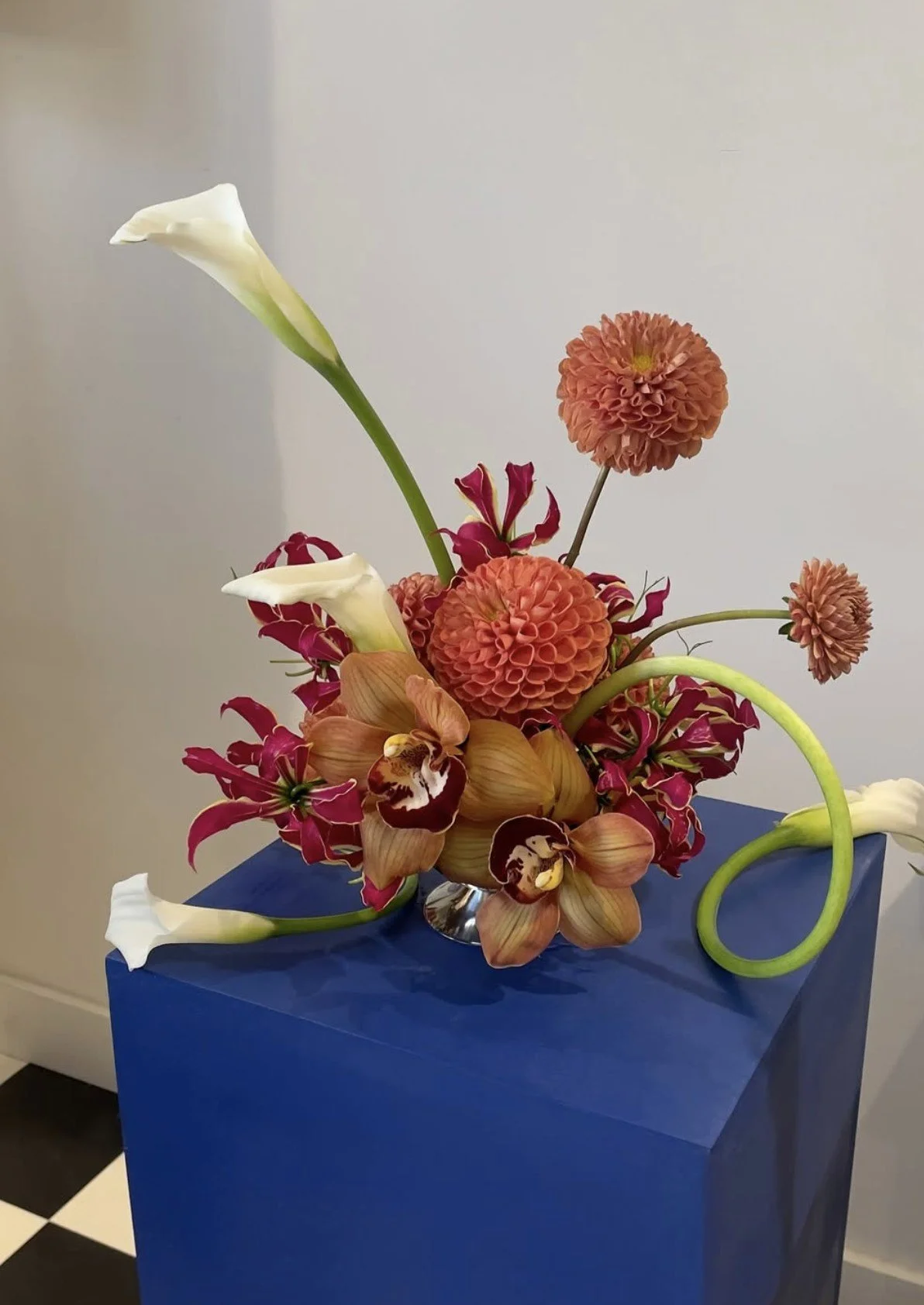 Composition florale avec calla lilies blanches, dahlias orange, orchidées cymbidium et lis gloriosa fuchsia, sur un socle bleu.